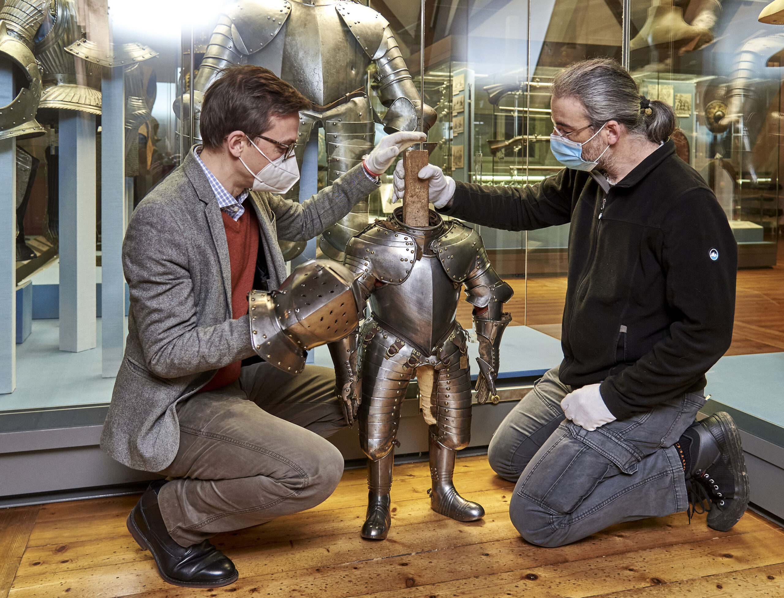 Two men wearing masks and gloves kneel beside a small suit of armor displayed in a museum at Hochschule Coburg. One adjusts the armor's shoulder piece while the other steadies it. A larger suit of armor looms in the background, with both men focused intently on the exhibit.