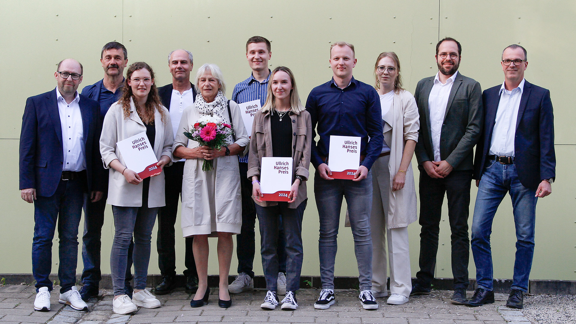 A group of 11 people stands proudly in front of a light-colored wall, representing Hochschule Coburg. Some hold awards, while the woman at the center cradles a bouquet of flowers. Dressed in semi-formal attire, they smile warmly at the camera.