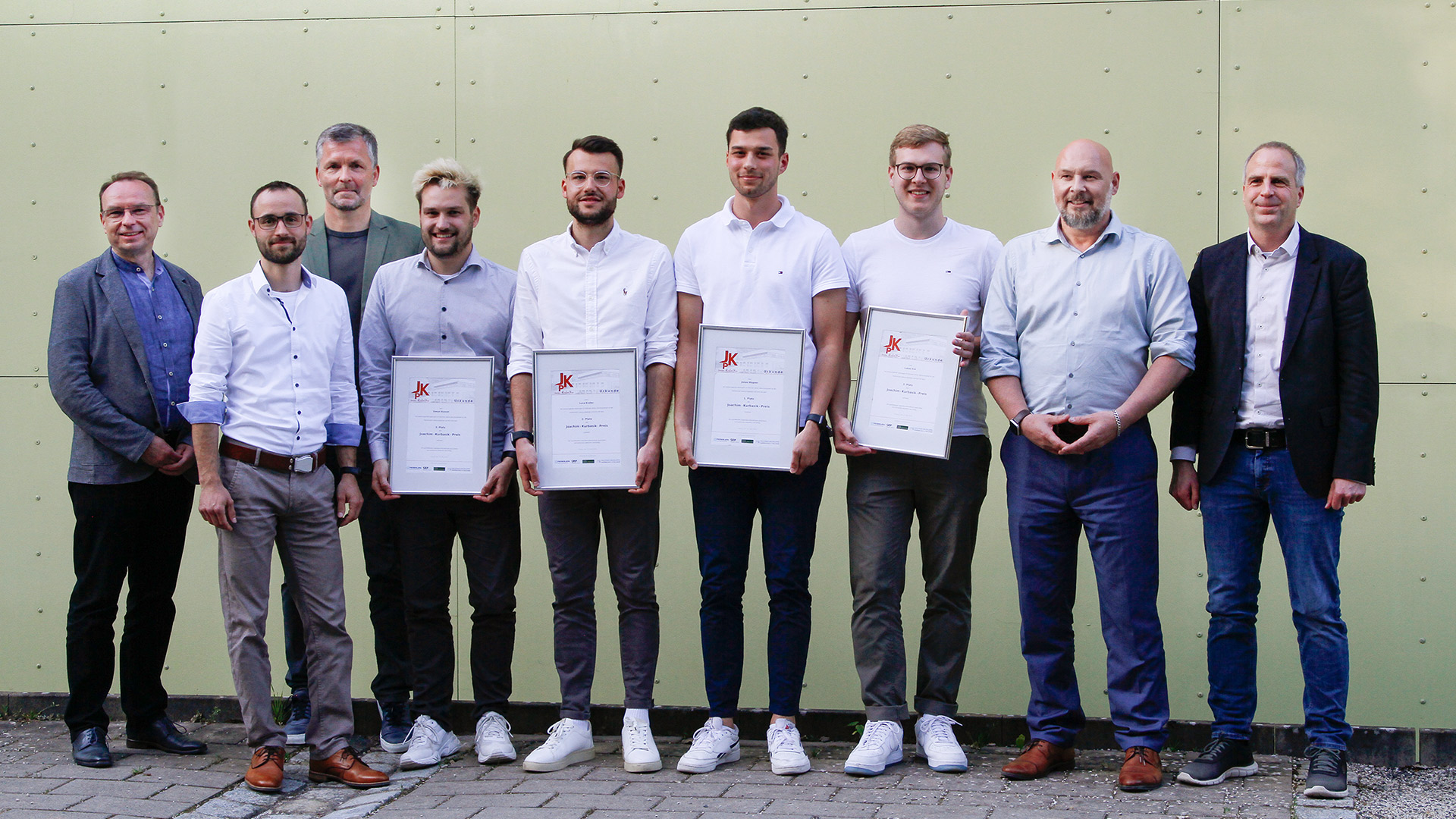 A group of nine people stands together in front of a light green wall at Hochschule Coburg. Four individuals in the middle hold certificates. Dressed in smart casual attire, everyone appears to be smiling, radiating a sense of achievement and camaraderie.