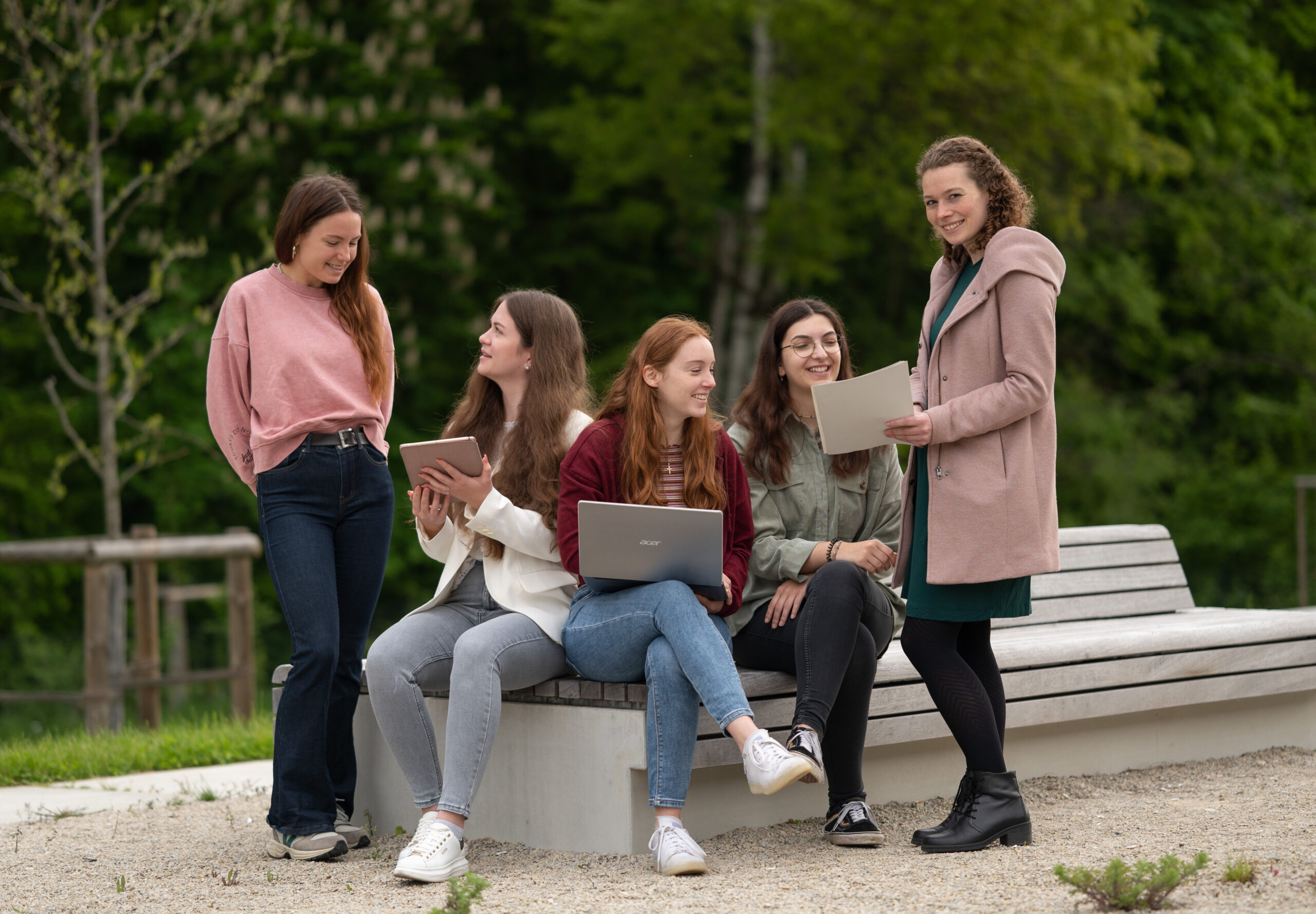 Fünf Frauen sitzen mit Laptops und Notizbüchern auf einer Bank im Freien an der Hochschule Coburg. Sie lächeln und unterhalten sich vor der üppig grünen Kulisse eines Parks.