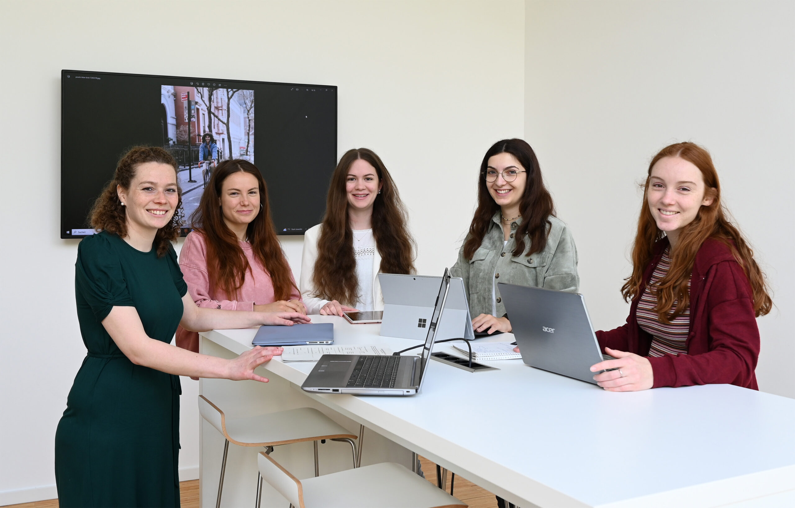 Fünf Personen stehen mit Laptops um einen weißen Tisch herum und lächeln in die Kamera. Im Hintergrund ist ein an der Wand montierter Bildschirm zu sehen, der ein Bild der Hochschule Coburg zeigt. Die Szenerie scheint ein Büro oder ein Besprechungsraum zu sein.