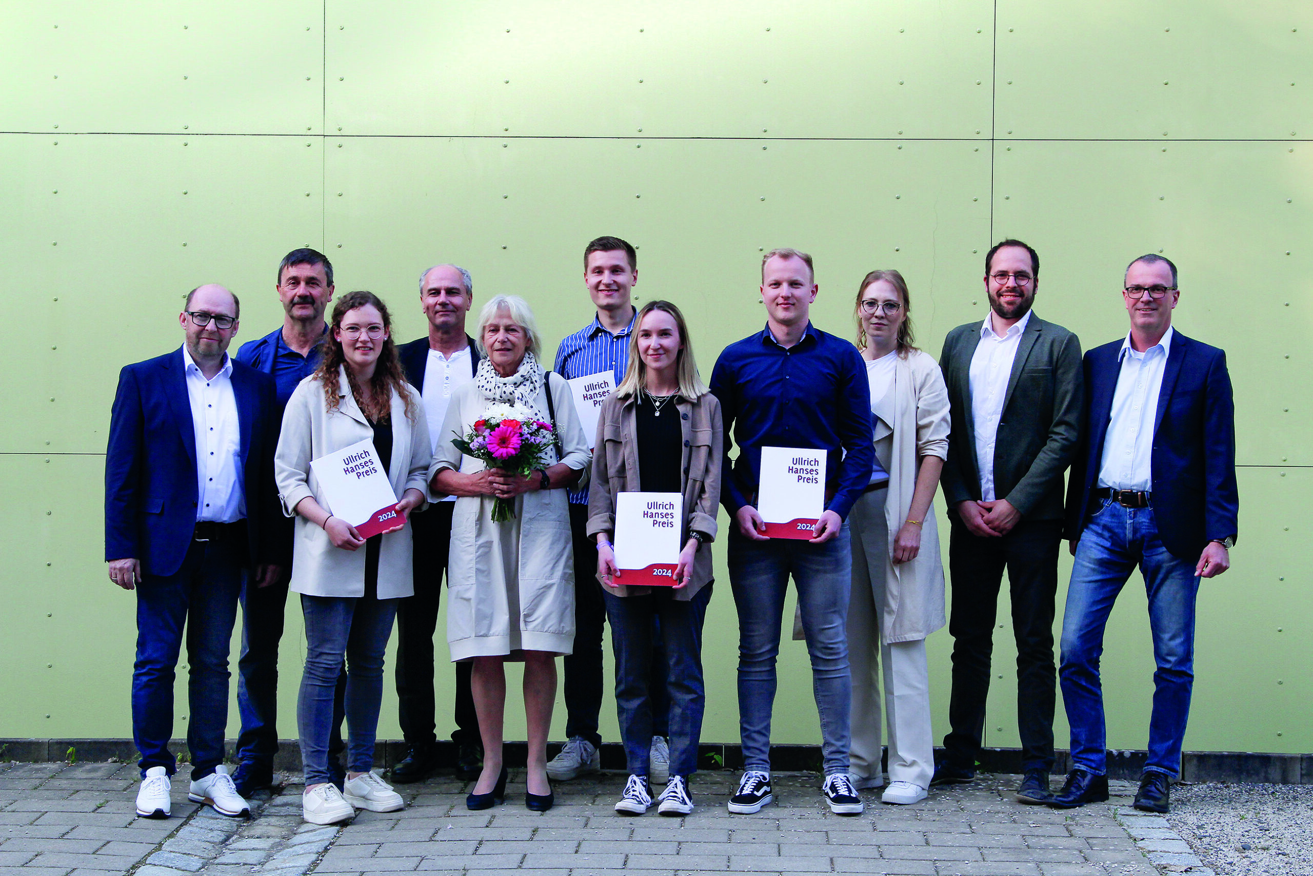 A group of eleven people stands in front of a light green wall at Hochschule Coburg. The woman in the center holds a bouquet of flowers, while four others proudly display certificates. Everyone is dressed in smart casual or business attire, capturing a moment of achievement and celebration.