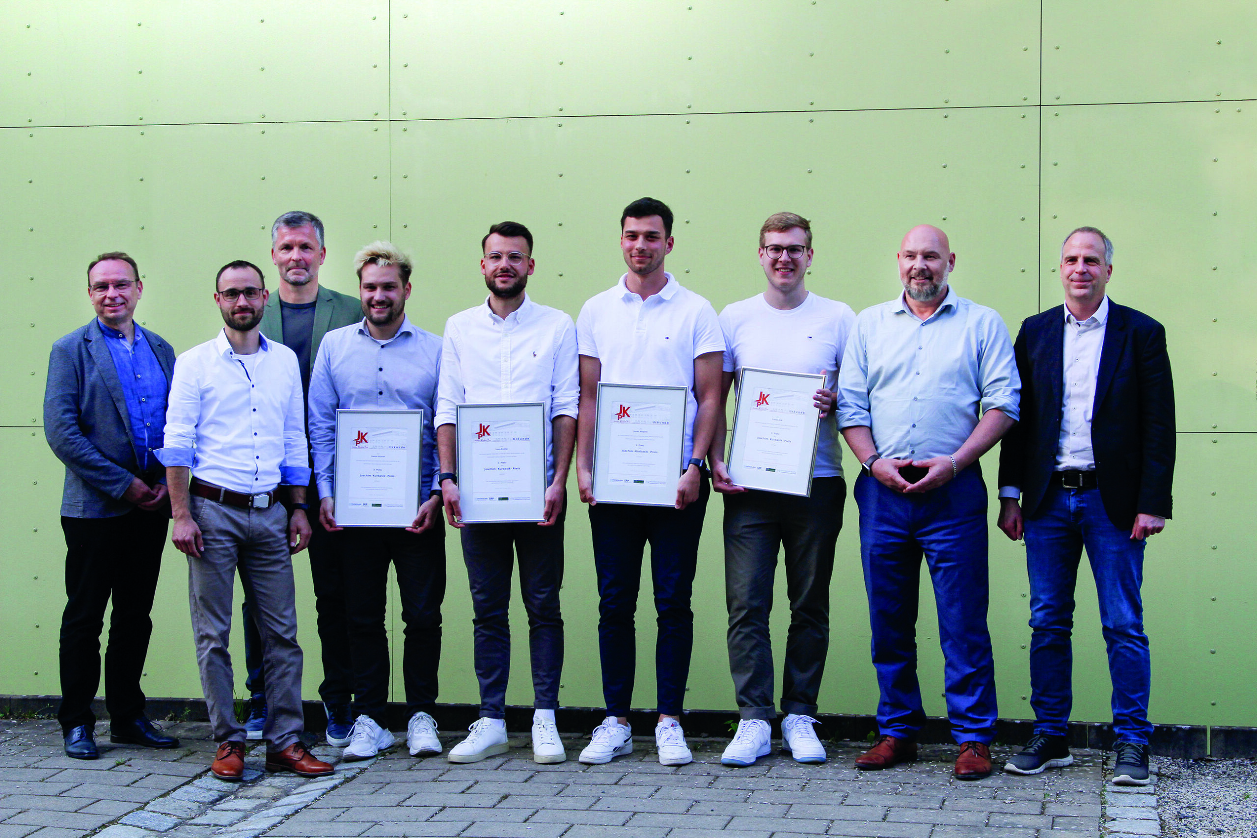 A group of nine men stand together in front of a green wall at Hochschule Coburg. Four men in the middle hold certificates, dressed in a mix of casual and business attire, as they pose for a group photo.