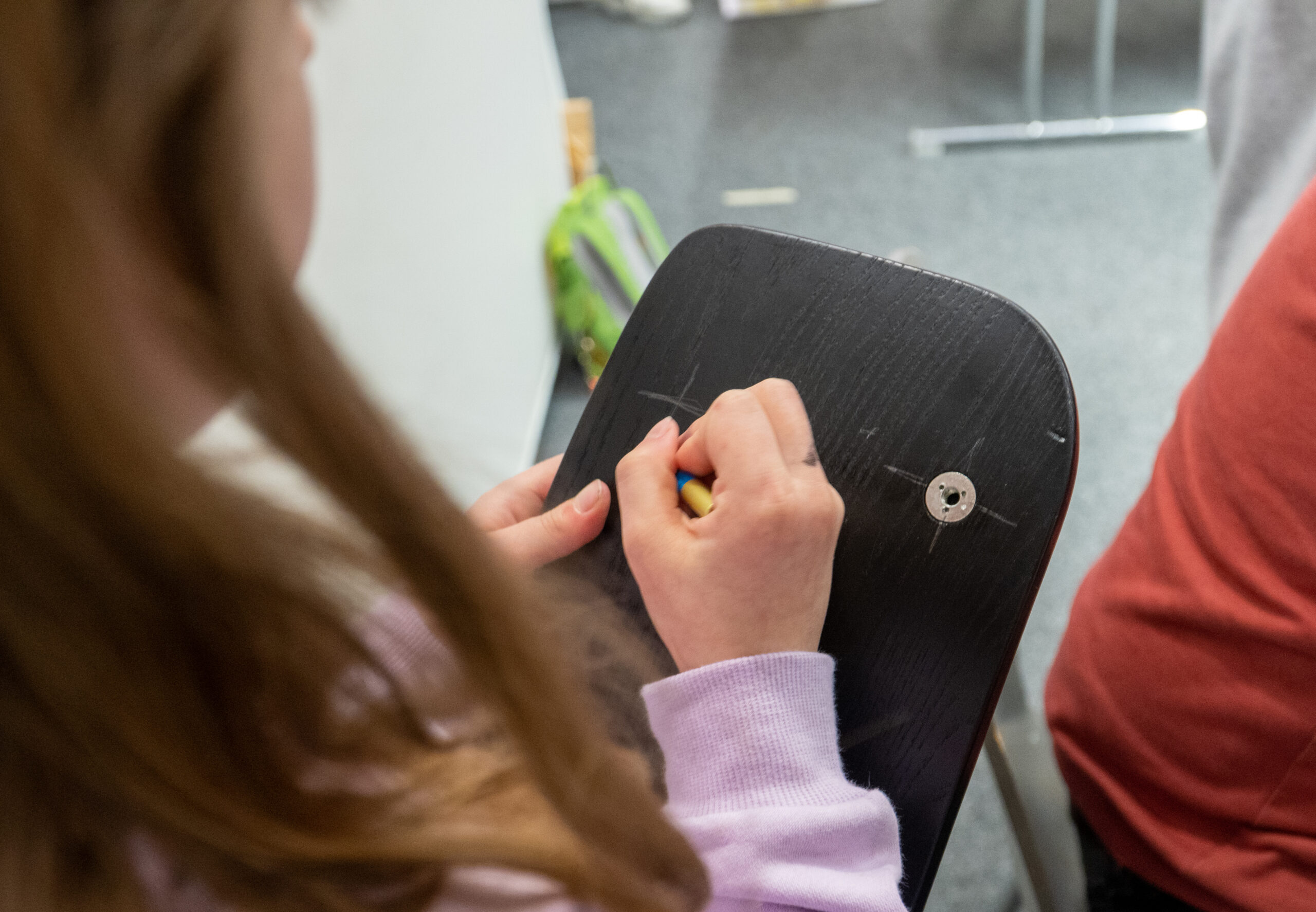 Eine Person mit langen Haaren und einem leichten Pullover sitzt auf einem Stuhl in der Hochschule Coburg und malt mit einem Buntstift ein kleines Muster auf die Rückenlehne eines anderen Stuhls. Die Szene scheint sich in einem Innenbereich zu befinden, der Boden ist mit grauem Teppich ausgelegt.