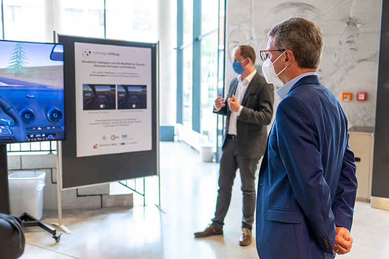 Ein Mann in Anzug und Maske beobachtet eine Präsentation über autonome Fahrtechnologie an der Hochschule Coburg. In der Nähe steht eine weitere maskierte Person neben einem Poster und einem Bildschirm, auf dem Fahrsimulationen gezeigt werden – alles in den großen Fenstern des modernen Gebäudes.