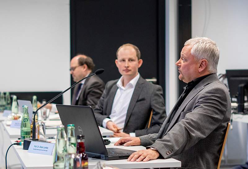 Drei Männer in Anzügen sitzen an einem Konferenztisch mit Mikrofonen, Laptops und Flaschen. Einer spricht, die anderen hören aufmerksam zu. Die Atmosphäre erinnert an ein formelles Meeting oder eine Präsentation an der Hochschule Coburg.