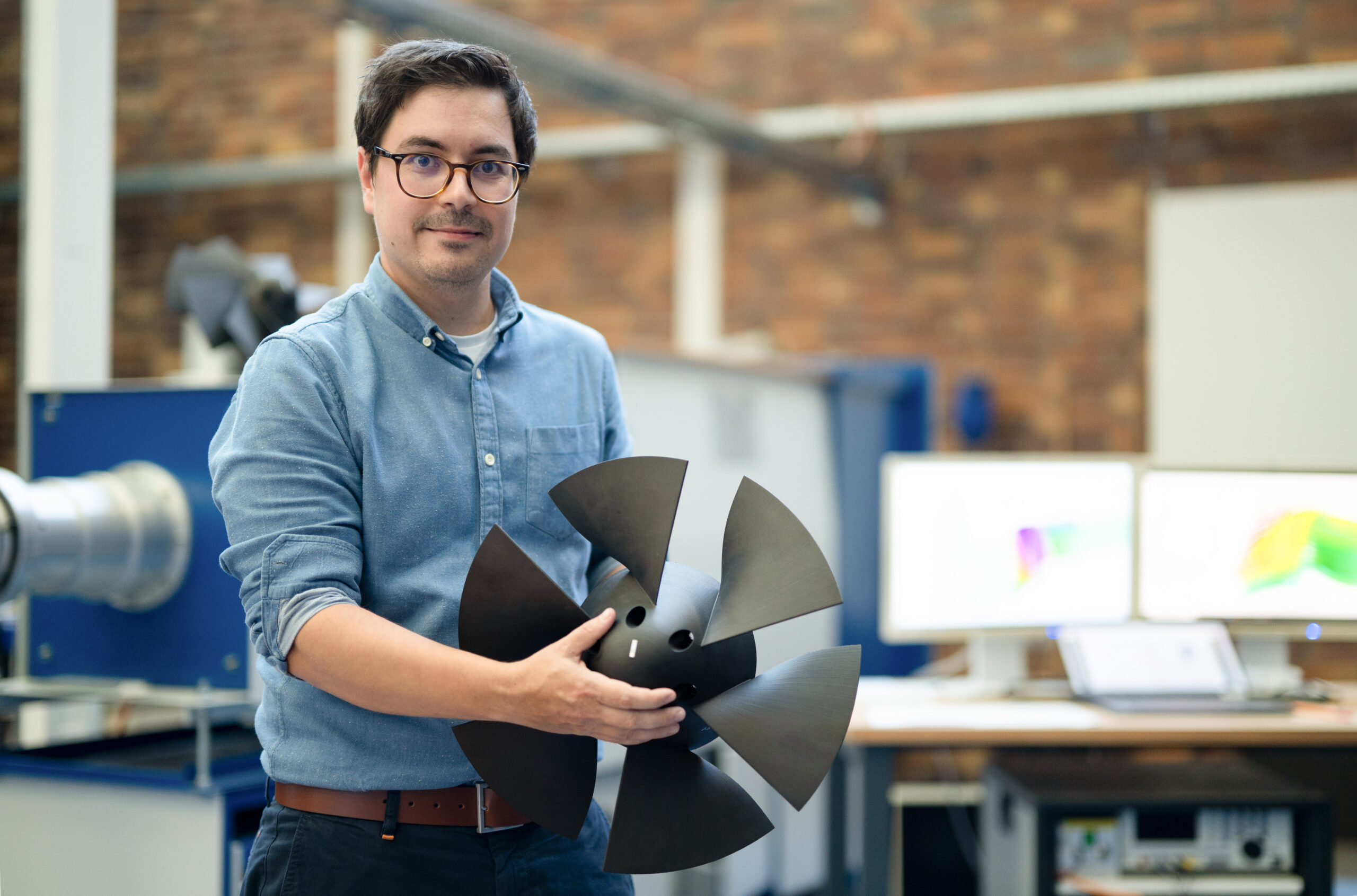 Eine Person mit Brille und blauem Hemd steht in einer Werkstatt der Hochschule Coburg und hält einen großen schwarzen Propeller in der Hand. Im Hintergrund zeigen Computerbildschirme bunte Grafiken und vor einer Backsteinwand stehen Industrieanlagen.