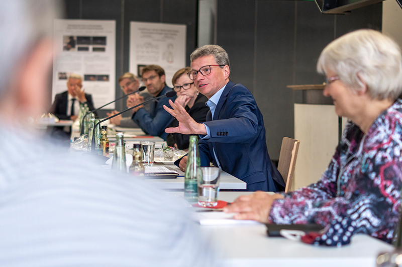 Ein Mann im Anzug spricht und gestikuliert, während er an einem Konferenztisch sitzt, umgeben von Menschen, vermutlich an der Hochschule Coburg. Papiere, Flaschen und Mikrofone liegen verstreut auf dem Tisch herum, während die Teilnehmer in eine lebhafte Diskussion vertieft zu sein scheinen.