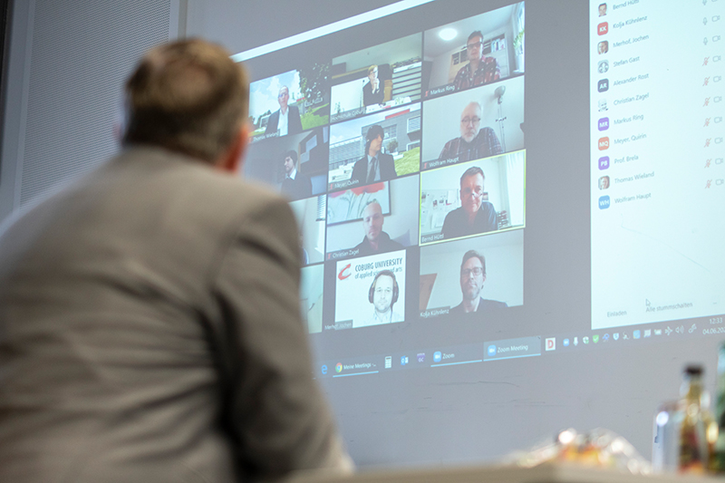 Eine Person im Anzug steht vor einem großen Bildschirm, auf dem eine Videokonferenz mit mehreren Teilnehmern läuft, was die vielfältige globale Reichweite der Hochschule Coburg veranschaulicht. Der Bildschirm zeigt eine Rasteransicht von Personen an verschiedenen Standorten, während im Vordergrund verschwommene Objekte auf dem Tisch liegen.