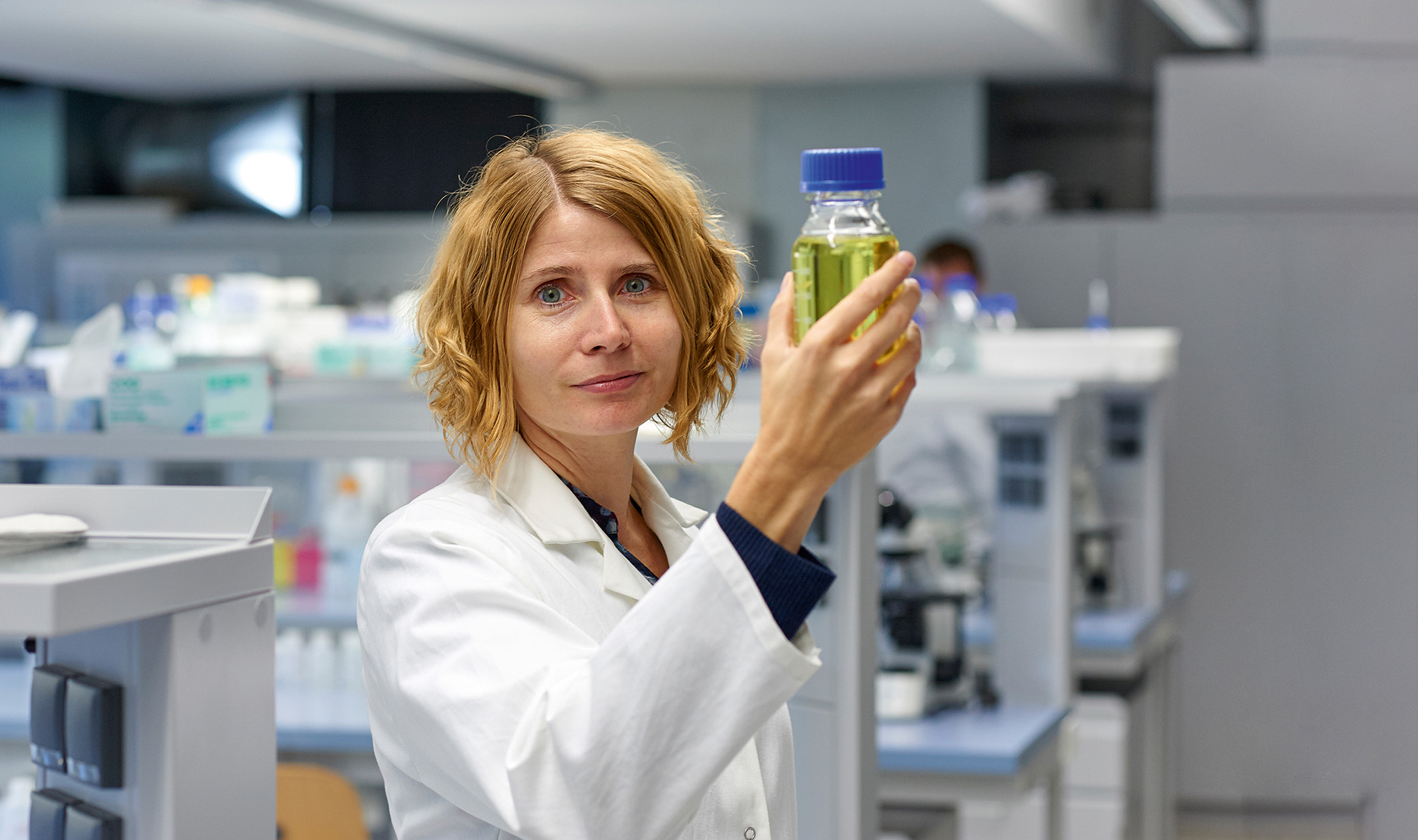 A woman in a laboratory coat holds up a bottle with yellow-green liquid, examining it closely. She stands in a modern laboratory at Hochschule Coburg, surrounded by various equipment in the background.