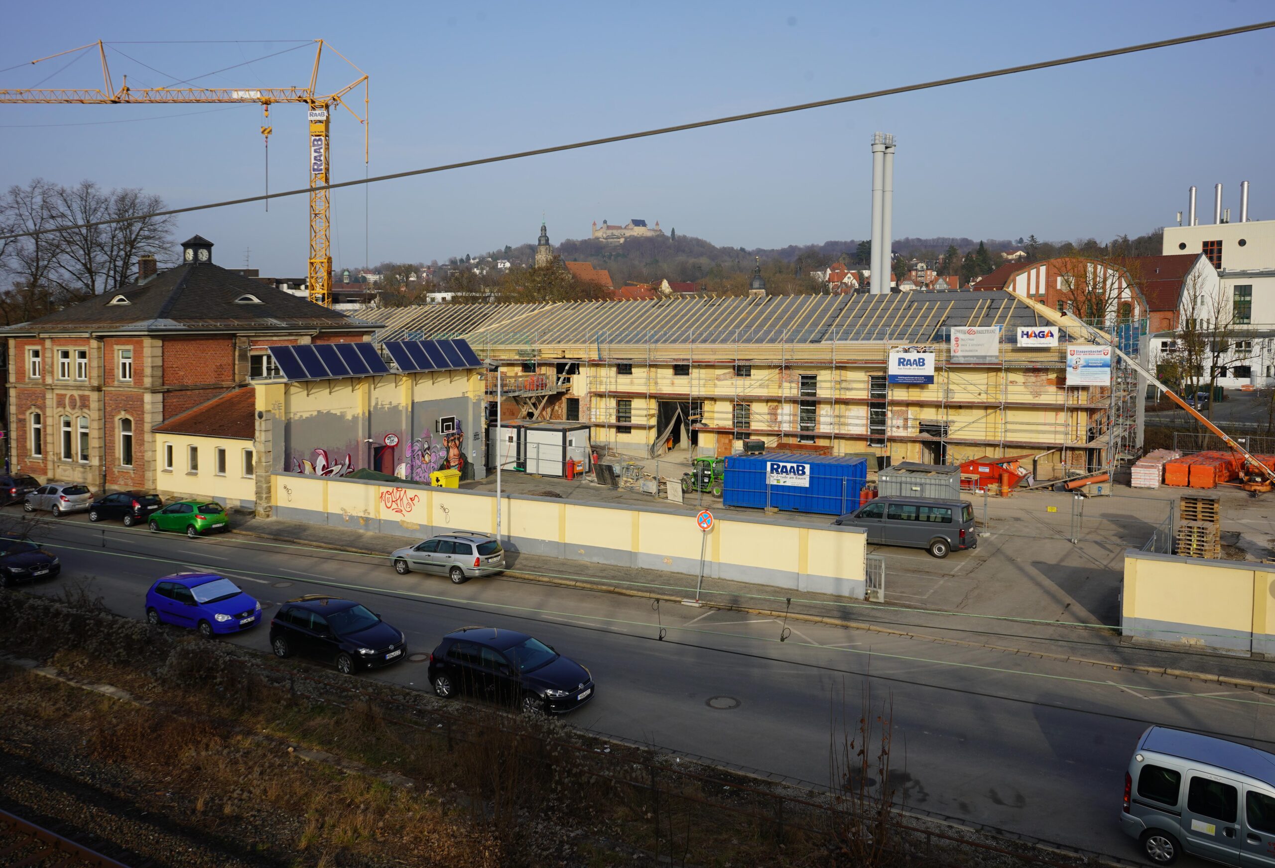 Straßenansicht einer Baustelle mit mehreren davor geparkten Fahrzeugen. In der Mitte steht ein großes Gebäude mit Gerüst, möglicherweise ein zukünftiger Flügel der Hochschule Coburg, mit einem Kran im Hintergrund. In der Ferne ist unter einem klaren Himmel eine Struktur auf einem Hügel zu erkennen.