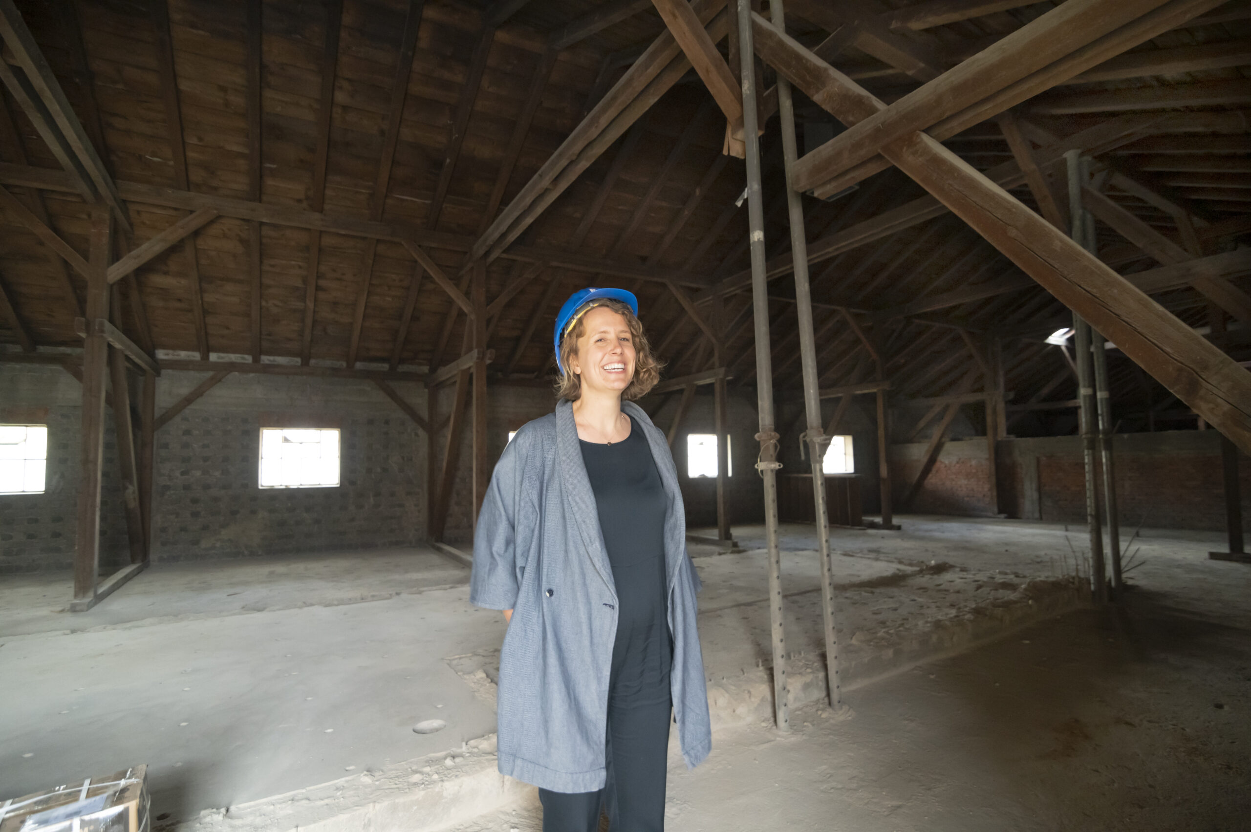 Eine Person mit blauem Bauhelm und grauem Mantel steht in einem großen, leeren Dachgeschoss mit Holzbalken und unfertigen Wänden. Lächelnd blickt sie von der Kamera weg in den Raum, der an der Hochschule Coburg renoviert wird.