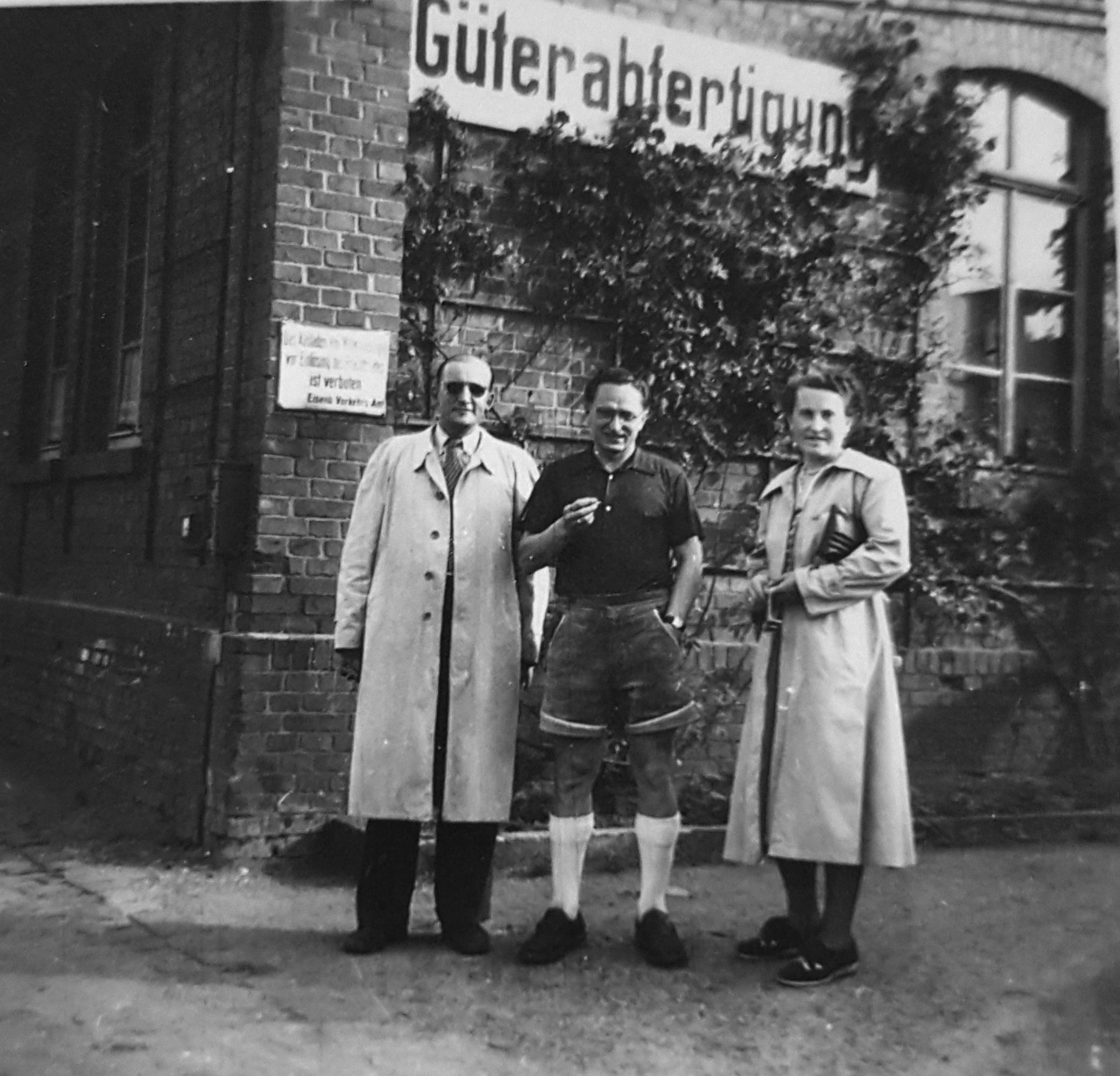 Drei Menschen stehen vor einem Backsteingebäude mit einem Schild mit der Aufschrift "Güterabfertigung". Zwei von ihnen tragen Trenchcoats, die Person in der Mitte trägt ein legeres Outfit, was darauf schließen lässt, dass es sich um Lehrkräfte der Hochschule Coburg handelt, die diesen Bahnhof oder das Lagergelände besuchen.