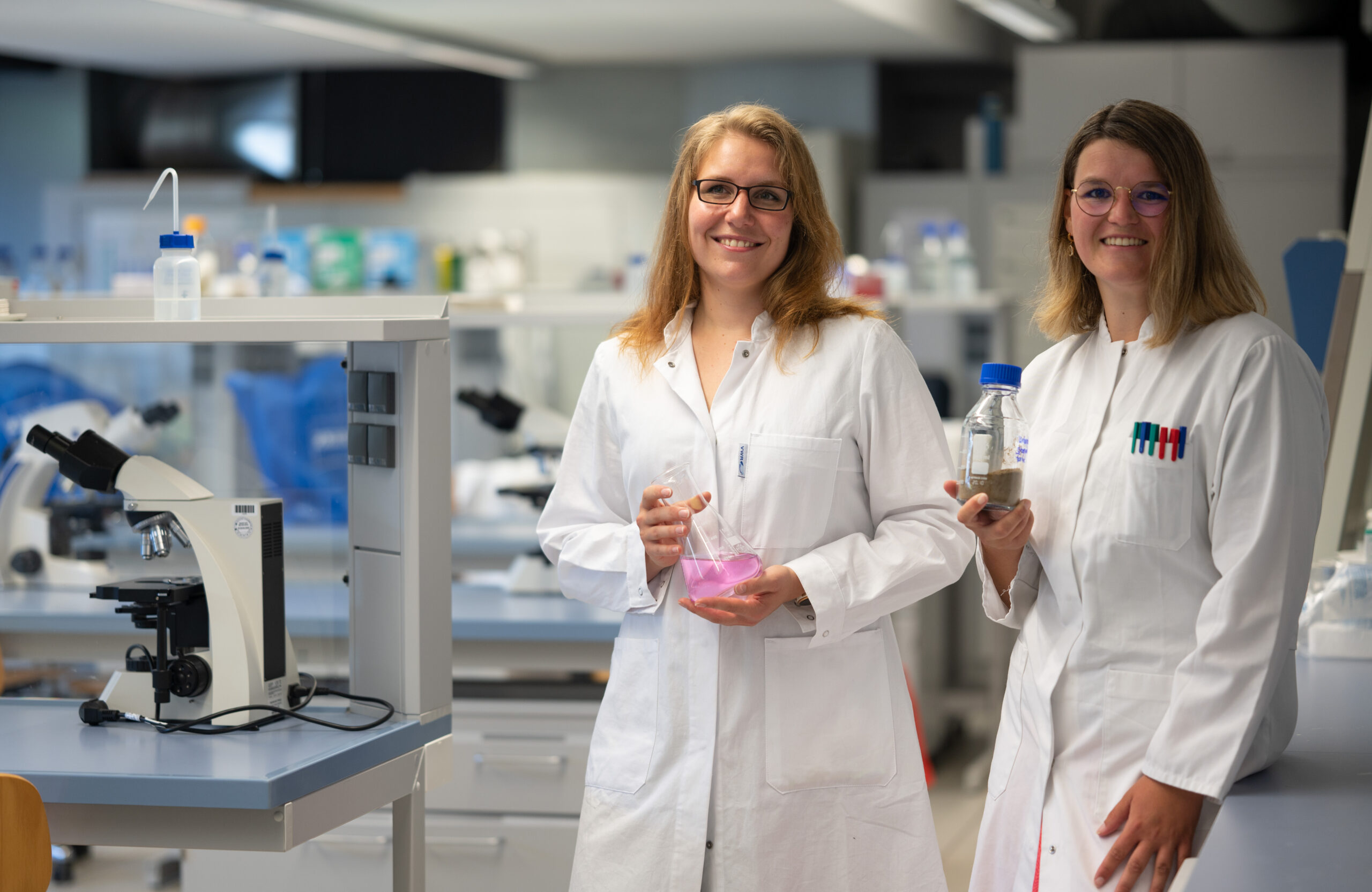 Zwei Wissenschaftler der Hochschule Coburg stehen lächelnd in Laborkitteln in einem modernen Labor. Einer hält einen Kolben mit rosa Flüssigkeit, der andere eine Flasche. Auf dem Tisch neben ihnen sind ein Mikroskop und elegante Laborgeräte zu sehen, die Innovation und Entdeckung symbolisieren.