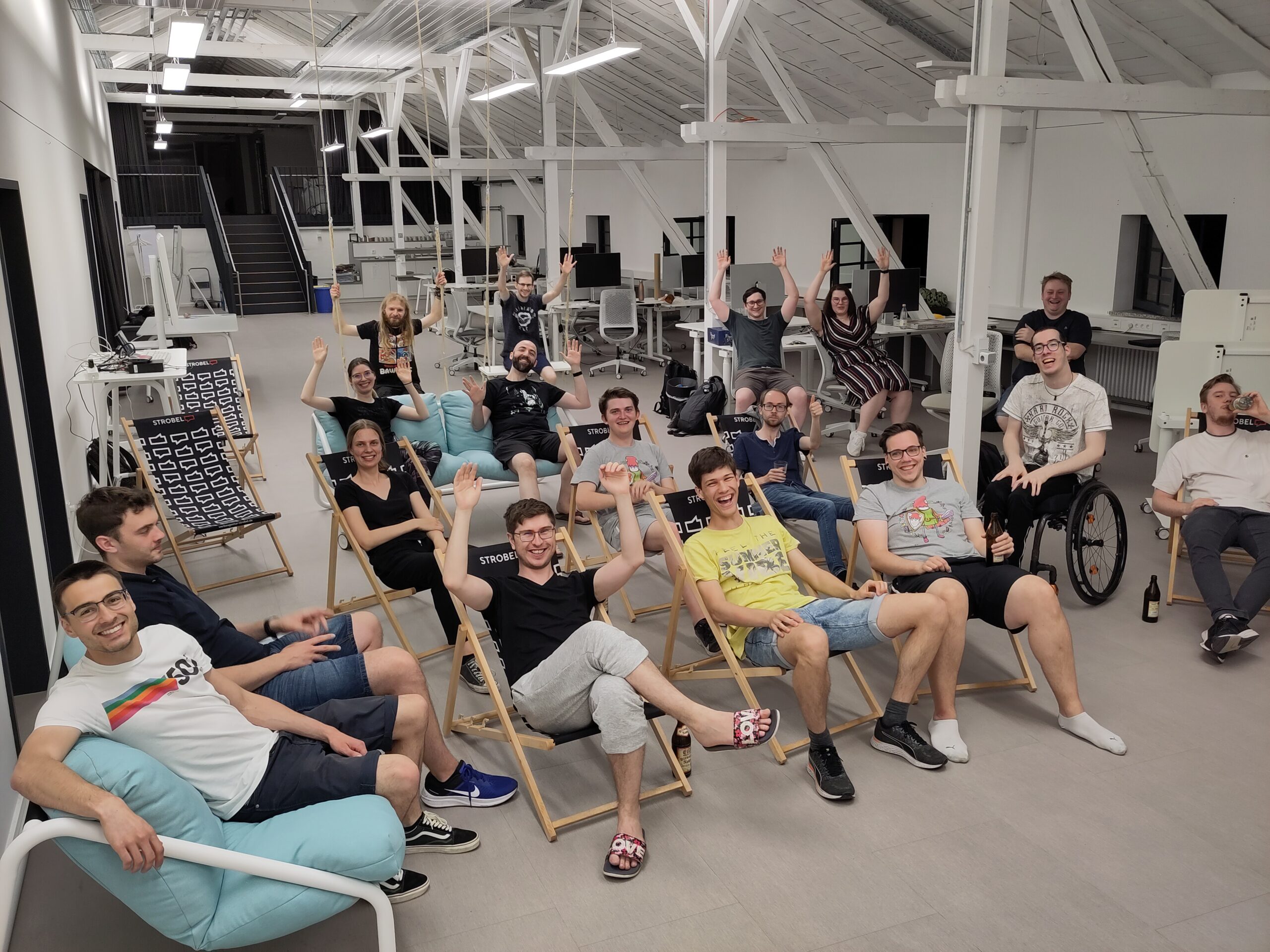 Eine Gruppe von Menschen der Hochschule Coburg sitzt in einem geräumigen, hellen Raum mit hoher Decke auf Liegestühlen und Sitzsäcken, lächelt und hebt die Hände. Einige benutzen Laptops, andere entspannen sich. Im Hintergrund sind Schreibtische und Bürogeräte zu sehen.