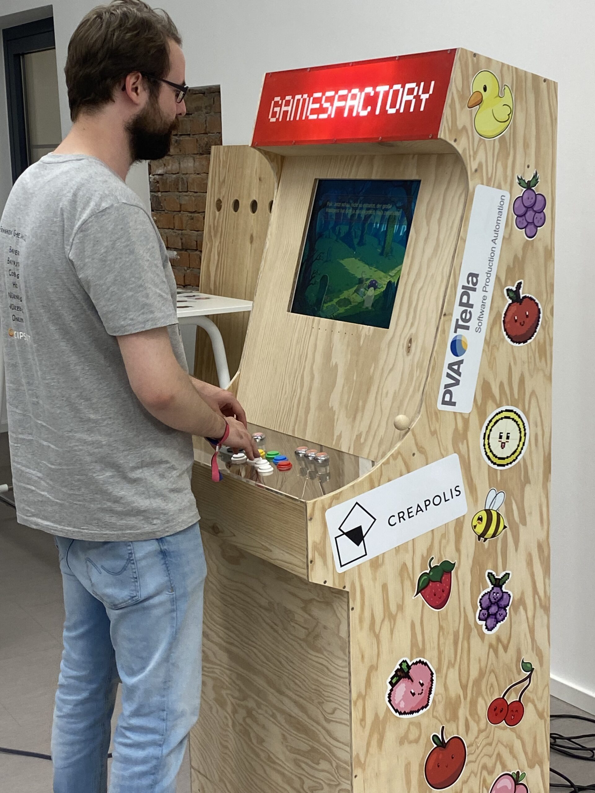 Eine Person ist in ein Retro-Arcade-Spiel vertieft, das auf einem Holzgehäuse mit einem „GAMESFACTORY“-Schild steht. Der Student der Hochschule Coburg trägt ein graues T-Shirt und Jeans, umgeben von Obstaufklebern und einer skurrilen Gummiente.