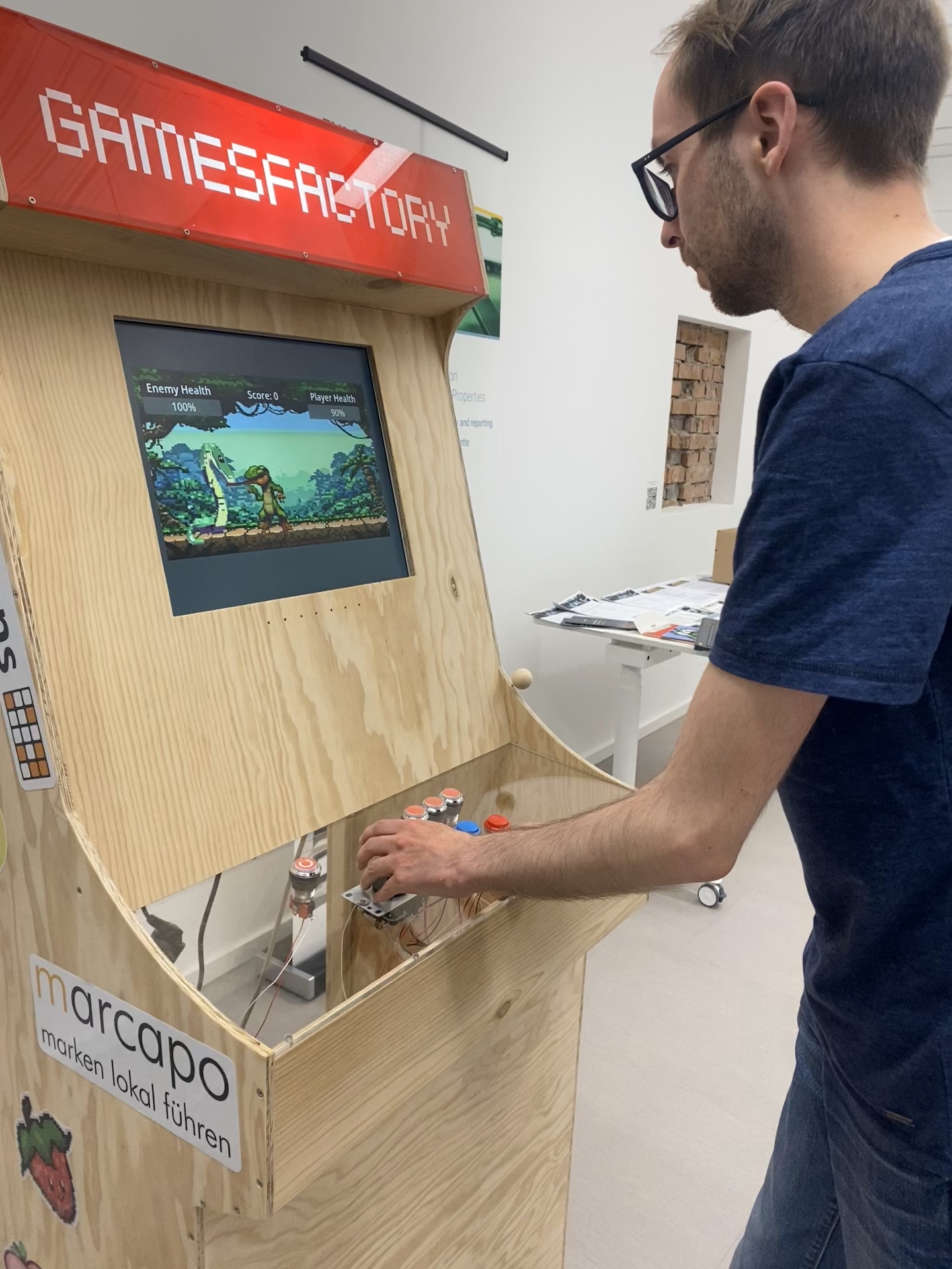 Eine Person ist in ein Videospiel vertieft, das auf einem hölzernen Arcade-Automaten mit der Aufschrift „GAMESFACTORY“ läuft. Das erinnert an die klassischen Studententreffs der Hochschule Coburg. Auf dem Bildschirm flimmert eine Kampfspielszene, umgeben von verschiedenen Knöpfen und einem Joystick. Auf der Vorderseite steht ein Aufkleber mit der Aufschrift „marcapo“.