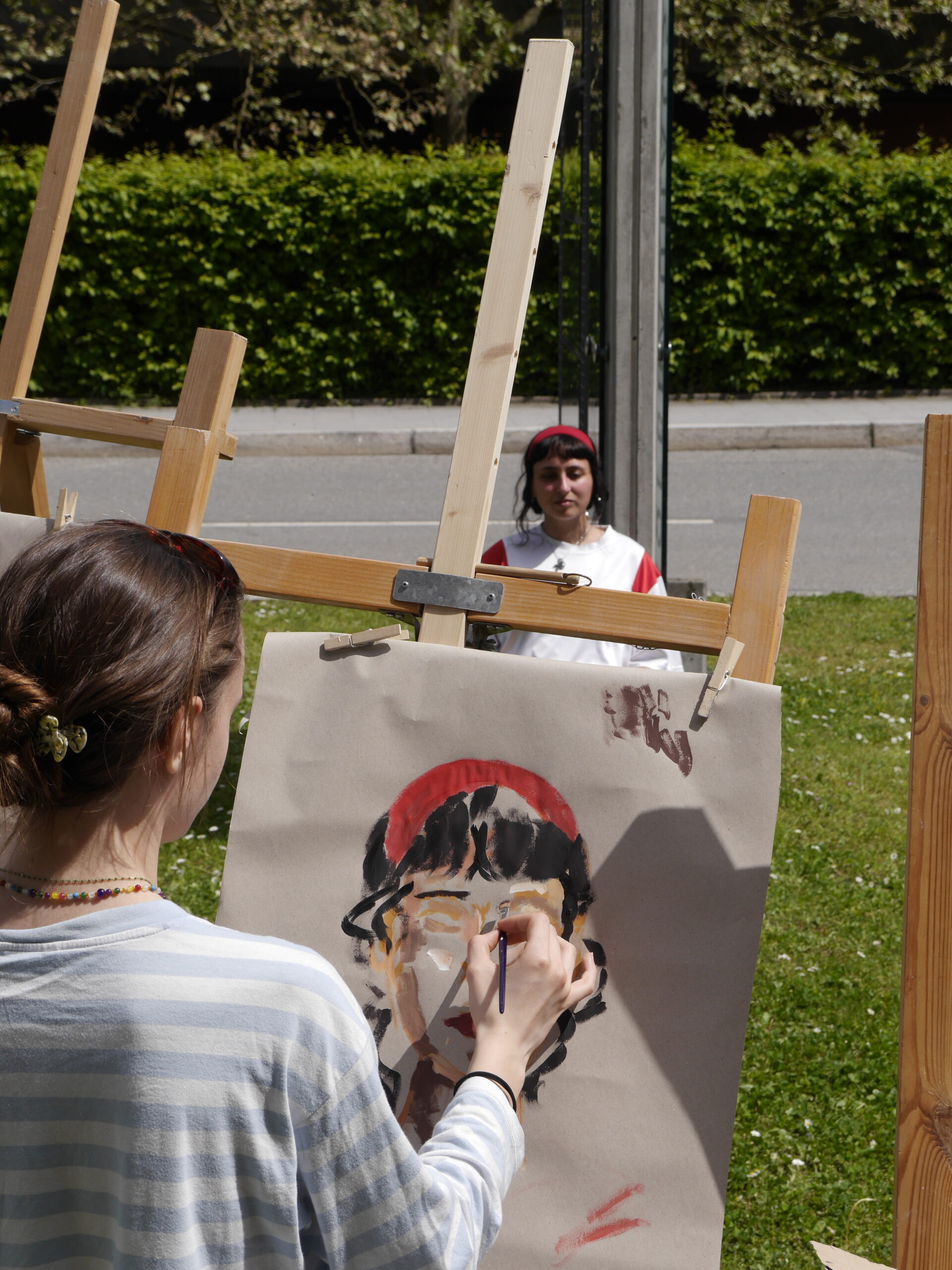 A person from Hochschule Coburg is painting a portrait outdoors on an easel, capturing another individual sitting in the background. The scene unfolds in a grassy area near a road with a hedge, where light and shadows highlight the creative process.