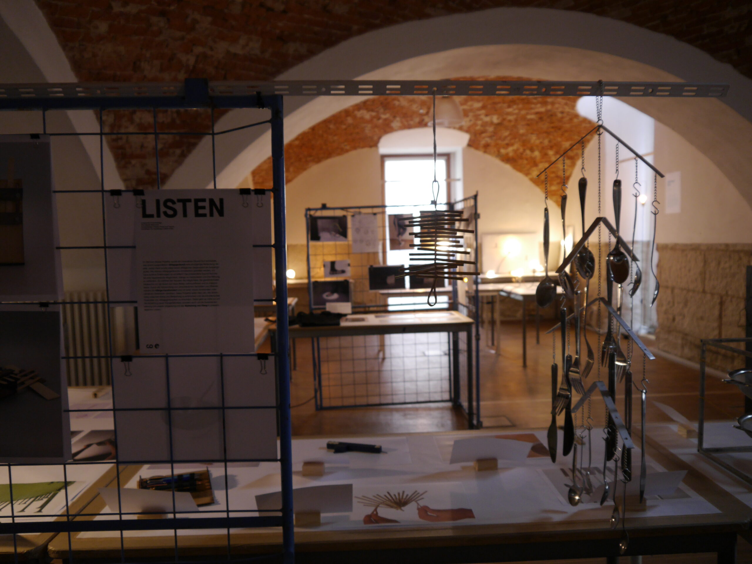 A dimly lit room with arched brick ceilings at Hochschule Coburg features an exhibition setup. Informational panels, hanging pans, and utensils are displayed, while tables showcase various papers and objects. The soft lighting creates a reflective atmosphere.