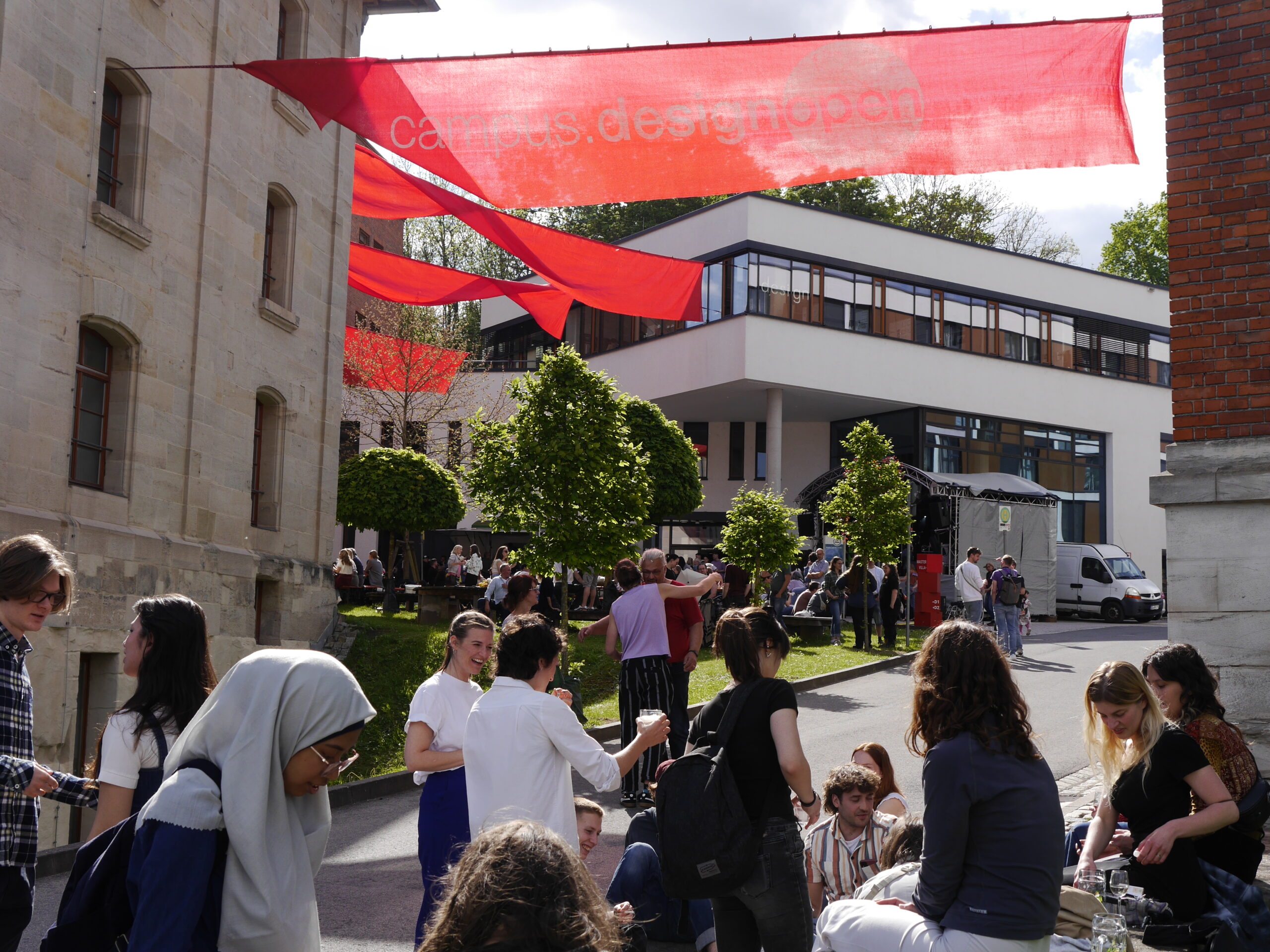 A lively outdoor gathering at Hochschule Coburg, with people socializing and eating on a sunny day. Red banners with white text are above, and a modern building along with trees provide the perfect backdrop.