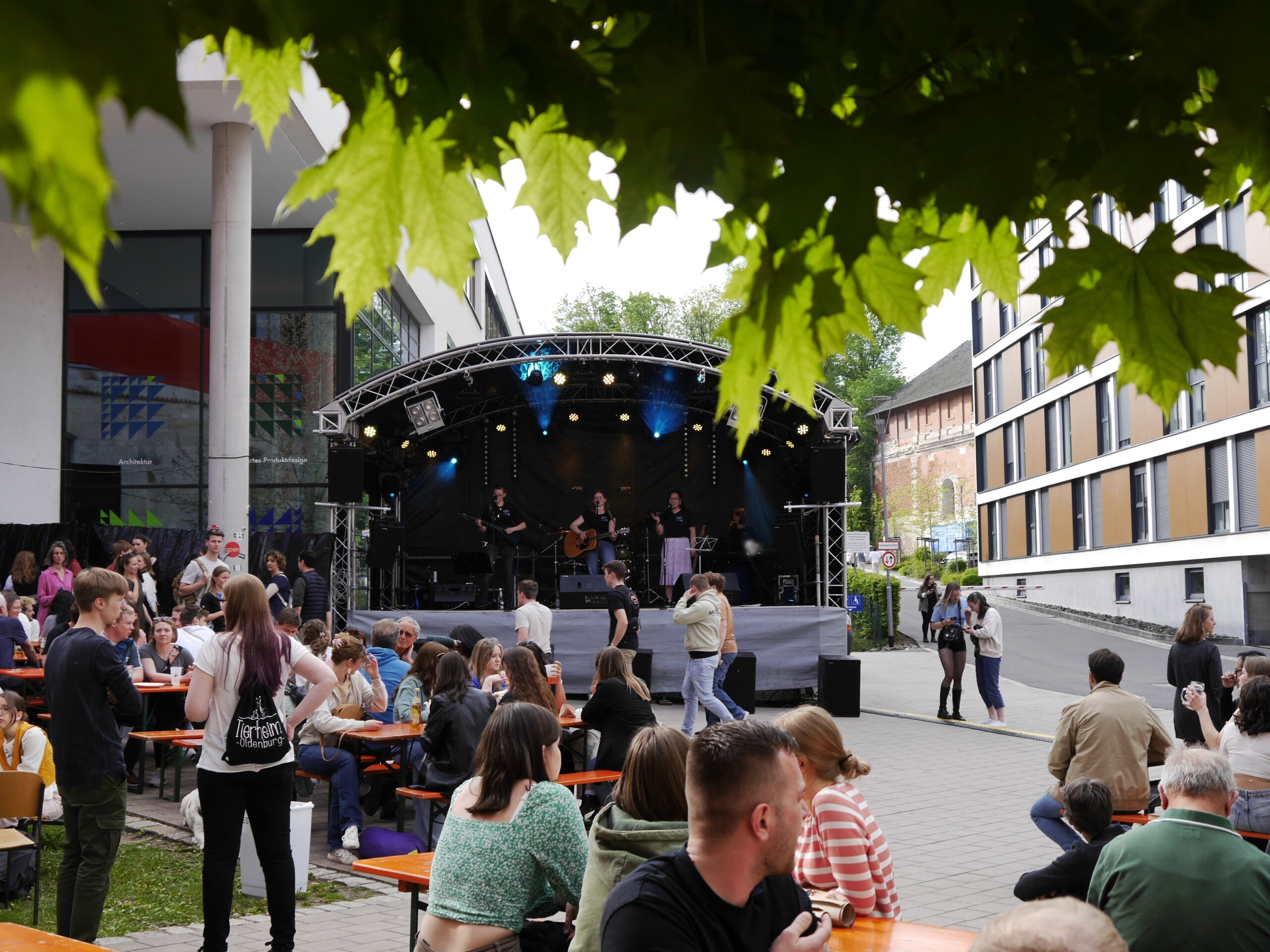 A lively outdoor concert near Hochschule Coburg features a band playing on stage under a canopy. People sit at picnic tables, enjoying the music. Green leaves frame the upper part of the image, adding a touch of nature to the urban setting.