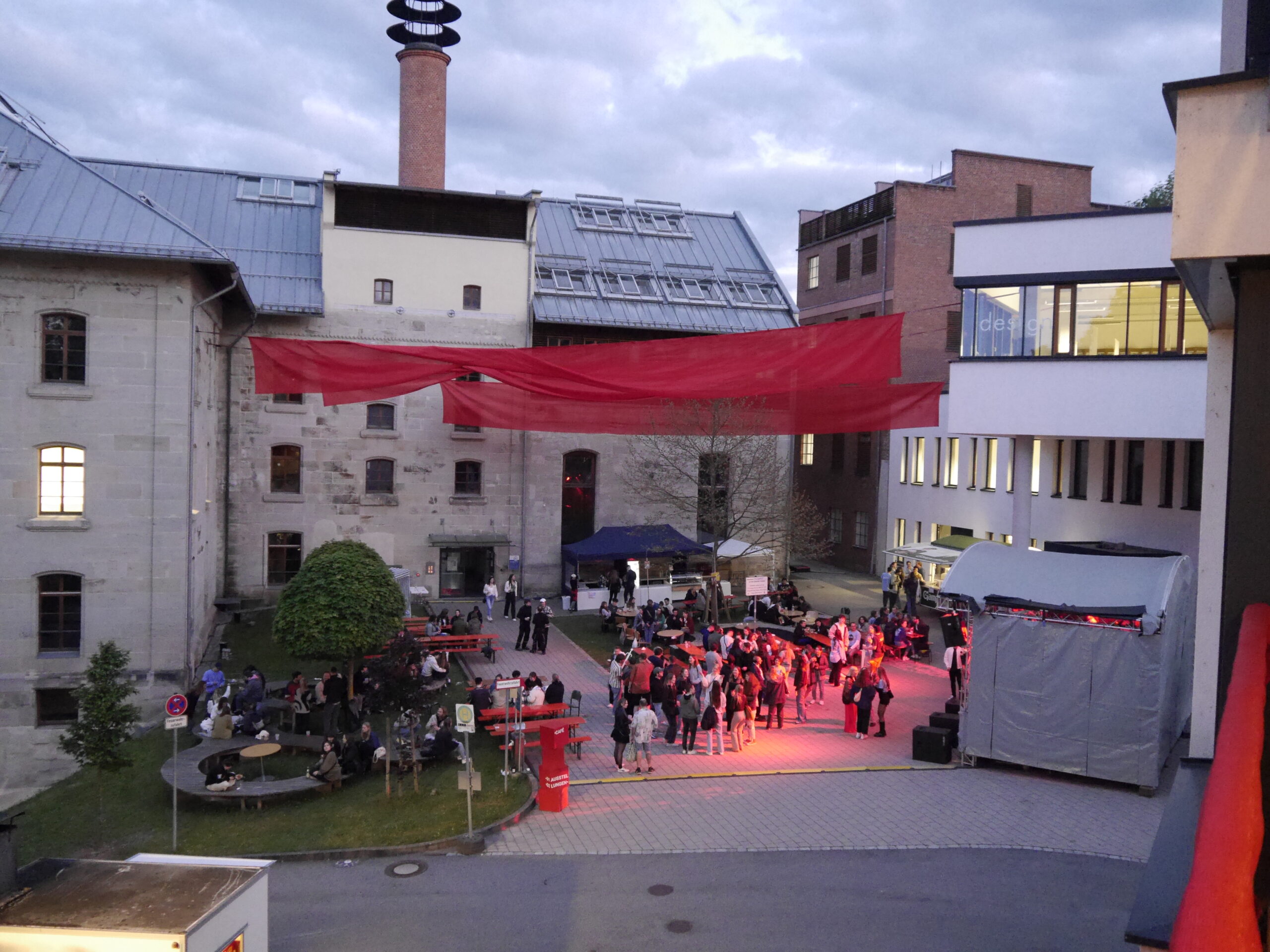 A lively courtyard gathering at Hochschule Coburg with people socializing and a small stage setup. Red fabric is suspended overhead, and buildings with a mix of modern and historic architecture surround the area. Evening lighting highlights the vibrant scene.