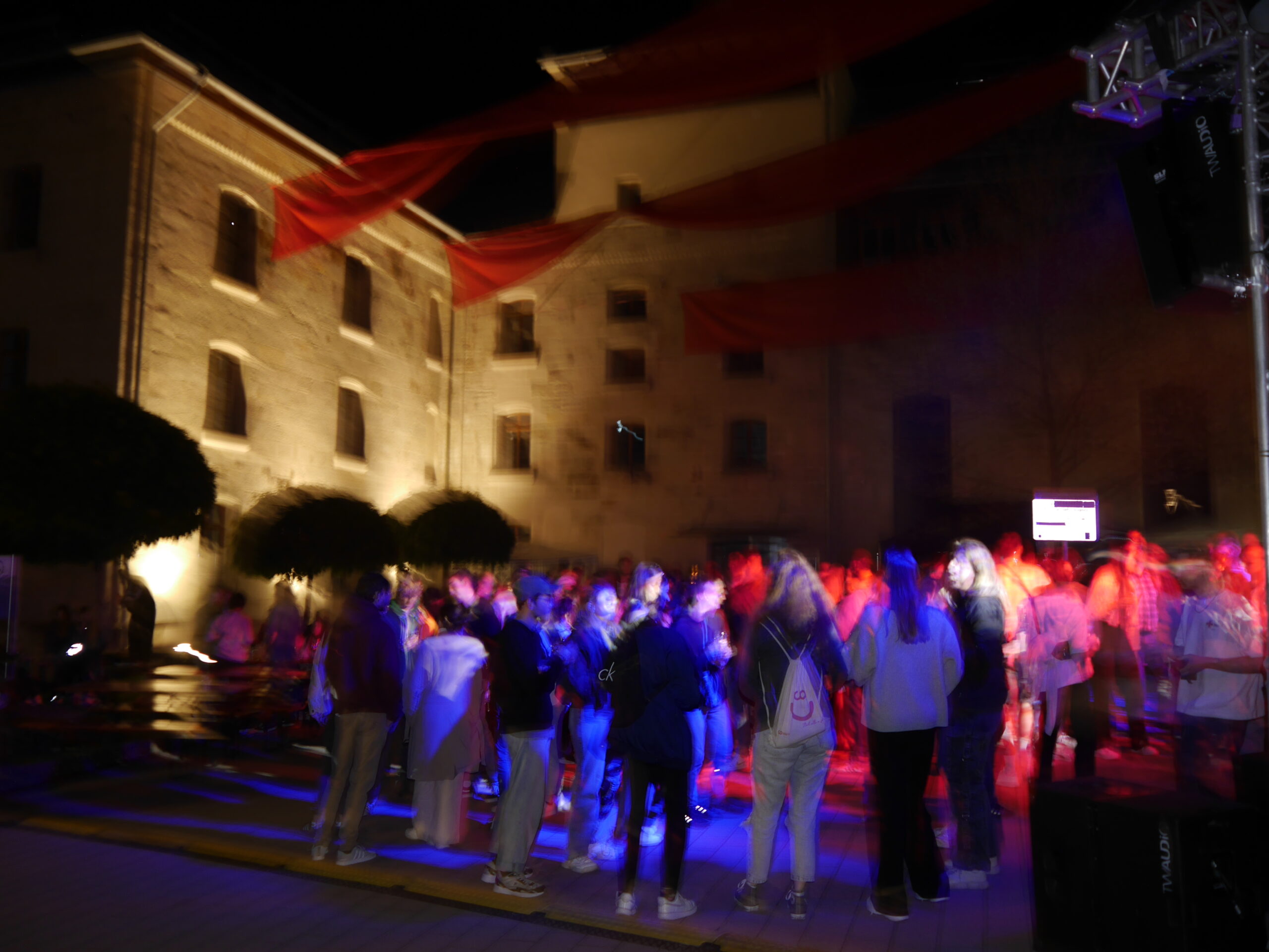 A lively crowd enjoys an outdoor night event at Hochschule Coburg, illuminated by colorful lights. Blurred figures suggest movement and dancing beneath red fabric decorations, with a large historic building serving as the enchanting backdrop.