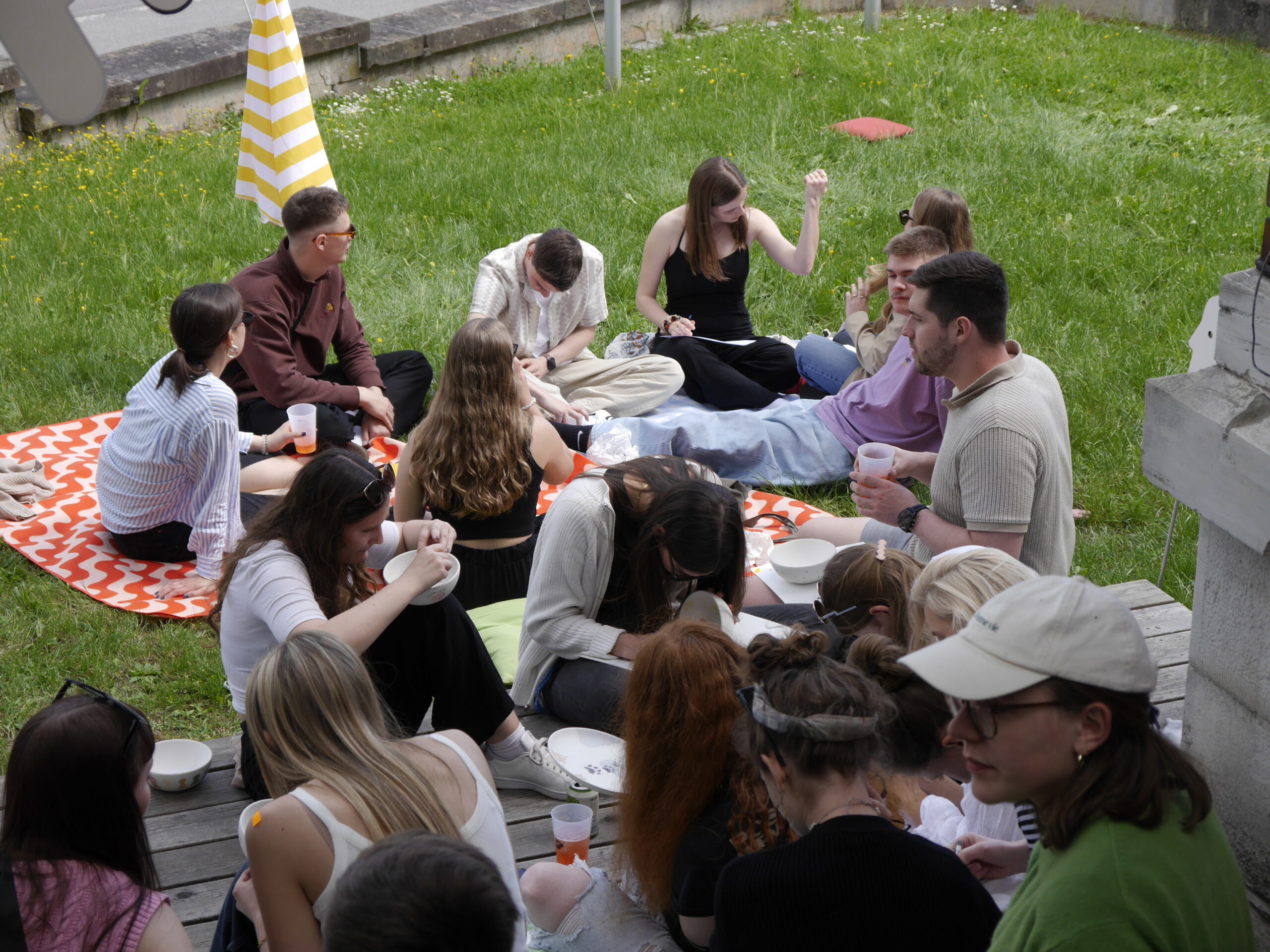 A group from Hochschule Coburg sits and relaxes on a wooden deck and grassy area during a casual outdoor gathering. Some are seated on blankets, holding drinks and engaging in conversation. A striped umbrella is partially visible in the background.