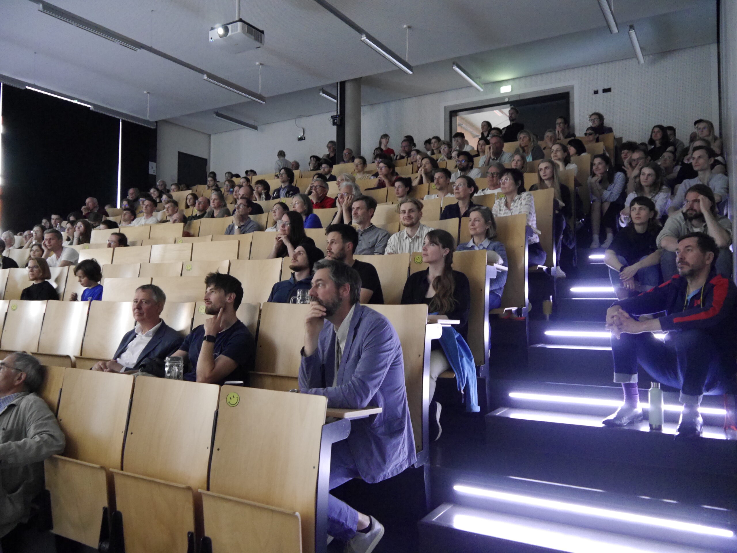 A large university lecture hall at Hochschule Coburg is filled with students seated in wooden chairs. They attentively watch a presentation, with some taking notes. The room is well-lit, and a projector is visible on the ceiling.