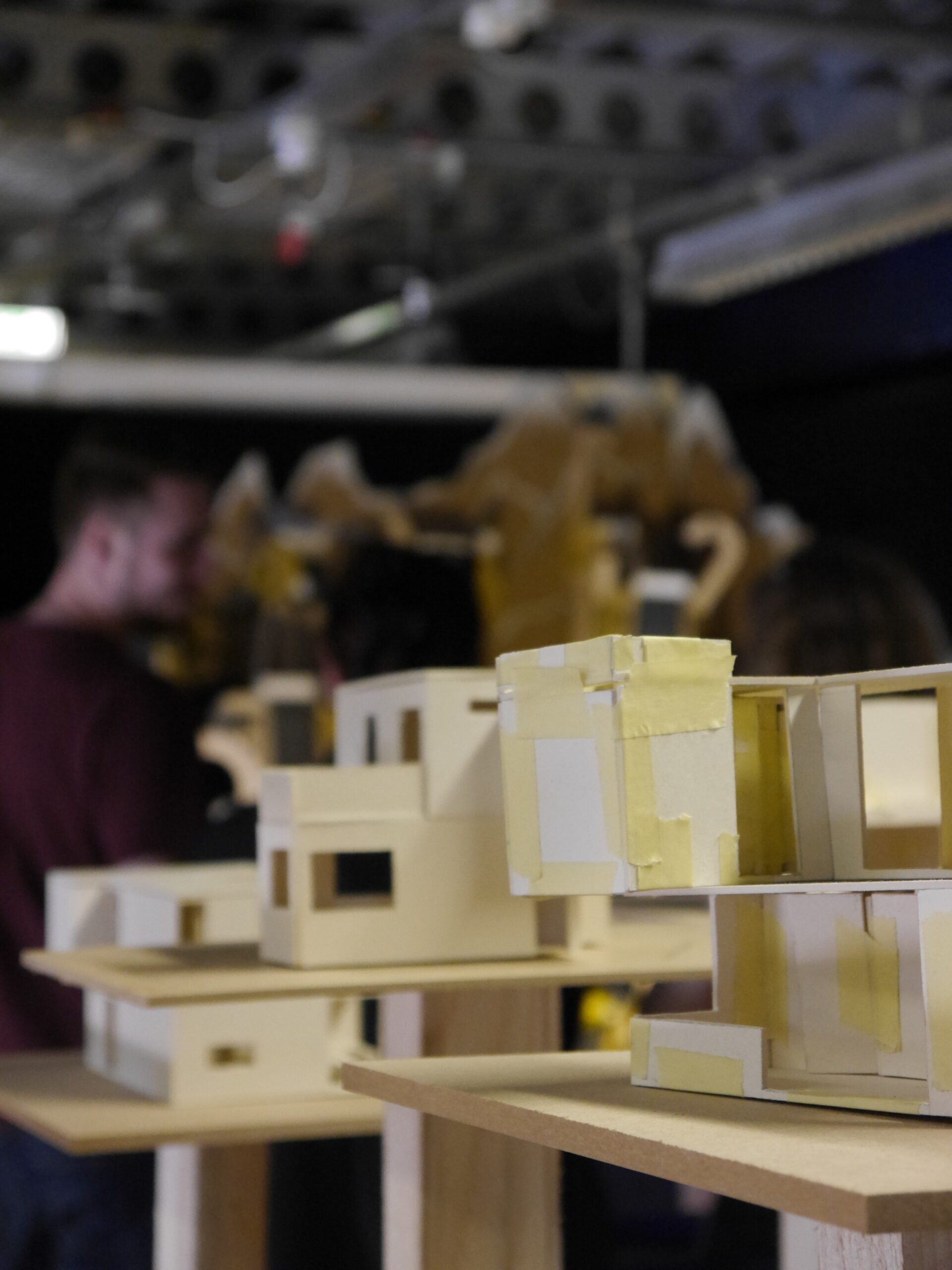 A close-up of architectural models made of cardboard with yellow tape, placed on wooden stands at a Hochschule Coburg event. In the background, people are blurred, enhancing the workshop or exhibition atmosphere.