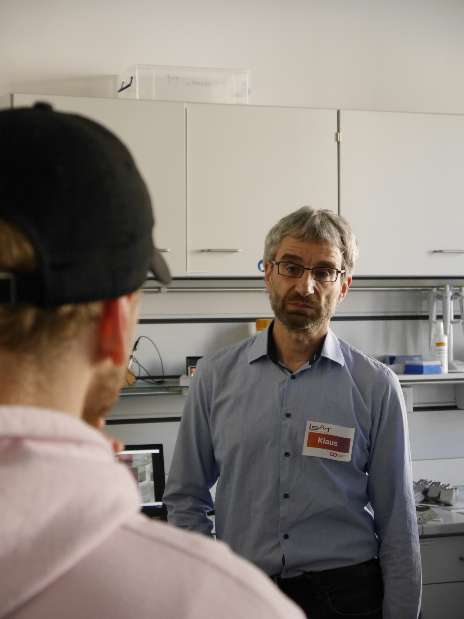 A man wearing a name tag stands in the laboratory of Hochschule Coburg, engaging in conversation with another person in a cap. The room features shelves, a computer, and various lab equipment in the background.