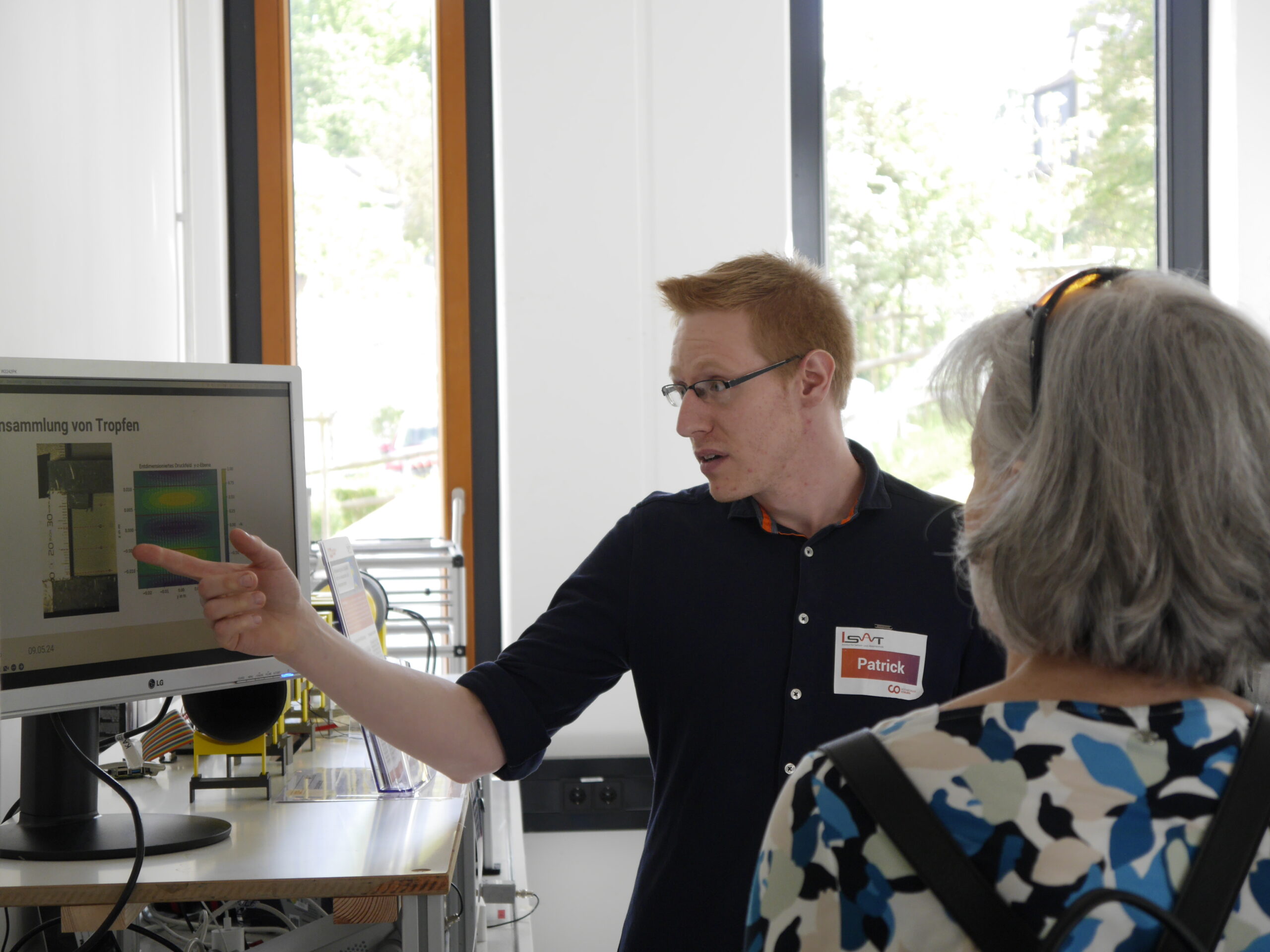 A person with a name tag "Patrick" is pointing at data on a computer screen, explaining to another person with gray hair. They are in a bright room with large windows and various electronic equipment, likely part of a cutting-edge project at Hochschule Coburg.