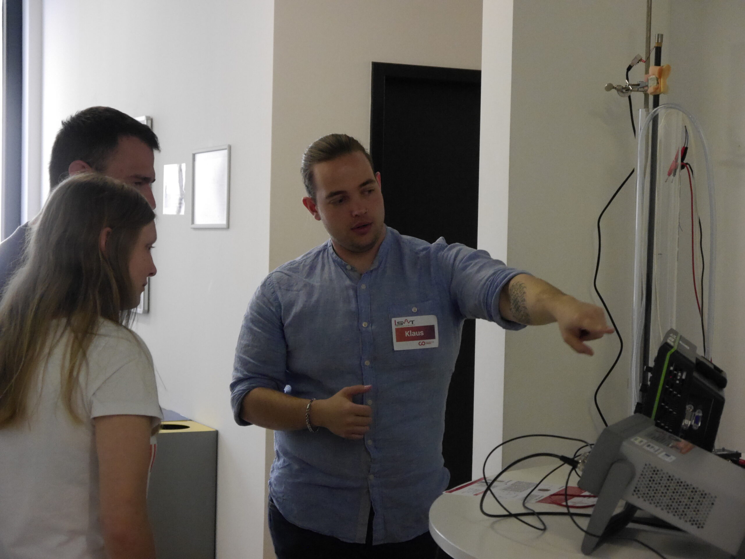 In an office setting at Hochschule Coburg, a person in a blue shirt is explaining something on a wired device to two others. The device sits on the table as the pair observes attentively.