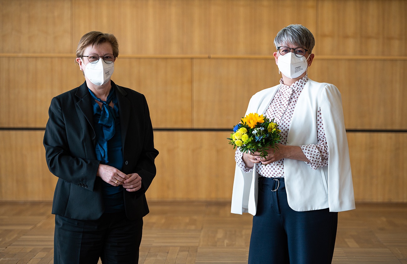 Zwei maskierte Personen stehen drinnen auf einem Holzboden in der Hochschule Coburg. Eine hält einen Strauß gelber und blauer Blumen in der Hand und trägt eine weiße Jacke und ein gepunktetes Hemd. Die andere trägt einen dunklen Anzug und eine blaugrüne Bluse. Beide haben kurze Haare und schauen in die Kamera.