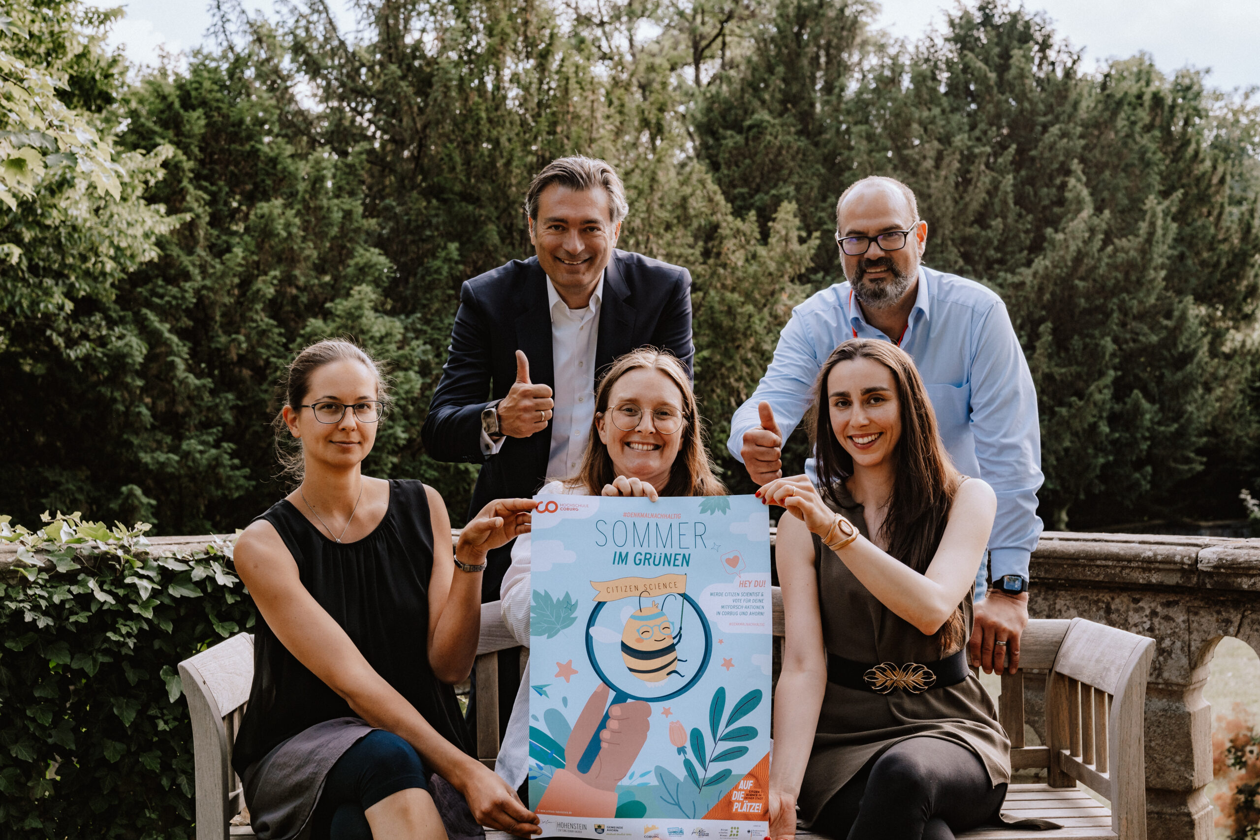 Fünf Menschen sitzen im Freien, lächeln und strecken den Daumen hoch. Sie halten ein buntes Plakat mit dem Text „Sommer im Grünen“ und einer Abbildung einer Katze in einem Blumentopf in den Händen. Im Hintergrund sind üppiges Grün und Bäume zu sehen, die den lebendigen Geist der Hochschule Coburg einfangen.