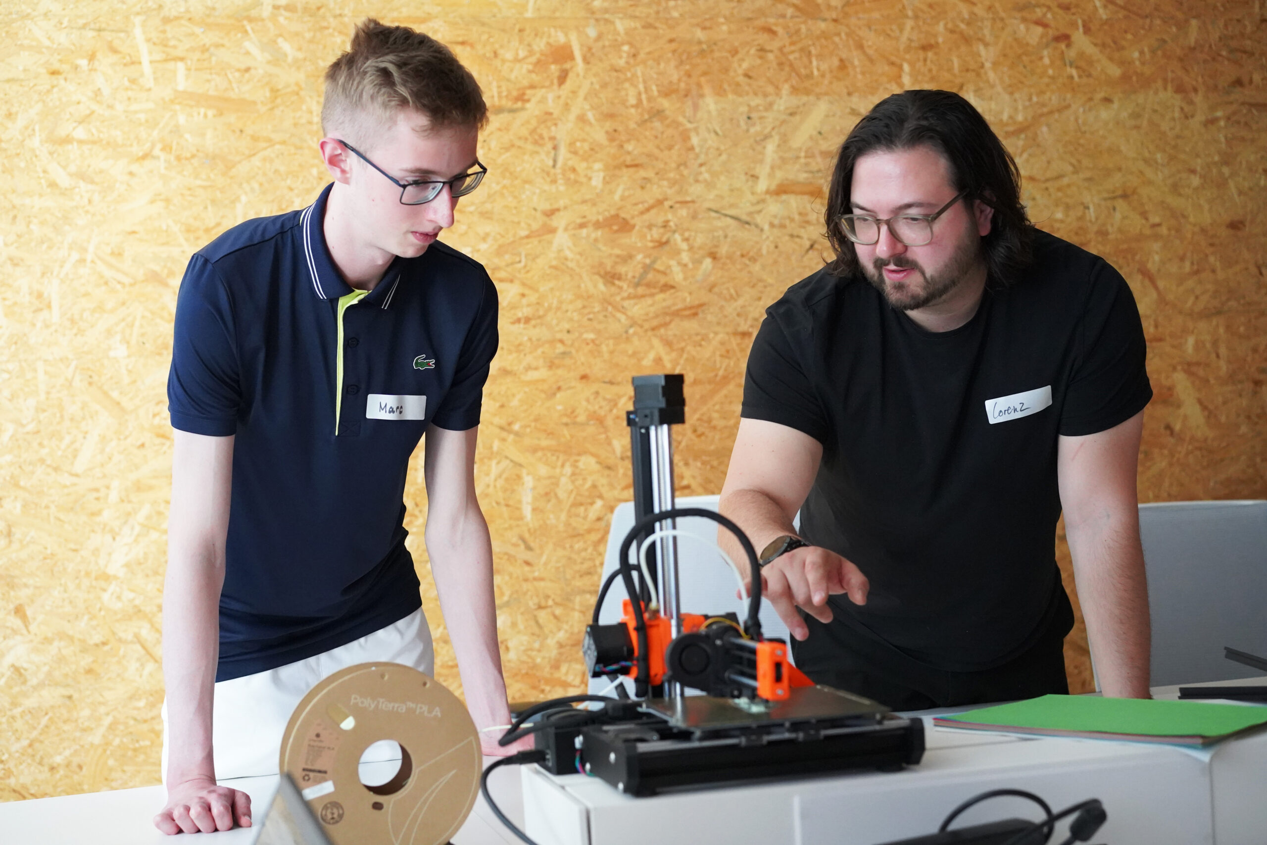 Two people, both with Hochschule Coburg name tags, are focused on a 3D printer on a table. One points at the printer while the other observes intently. A spool of filament and some papers are also visible against the textured wall background, adding to the scene's academic ambiance.