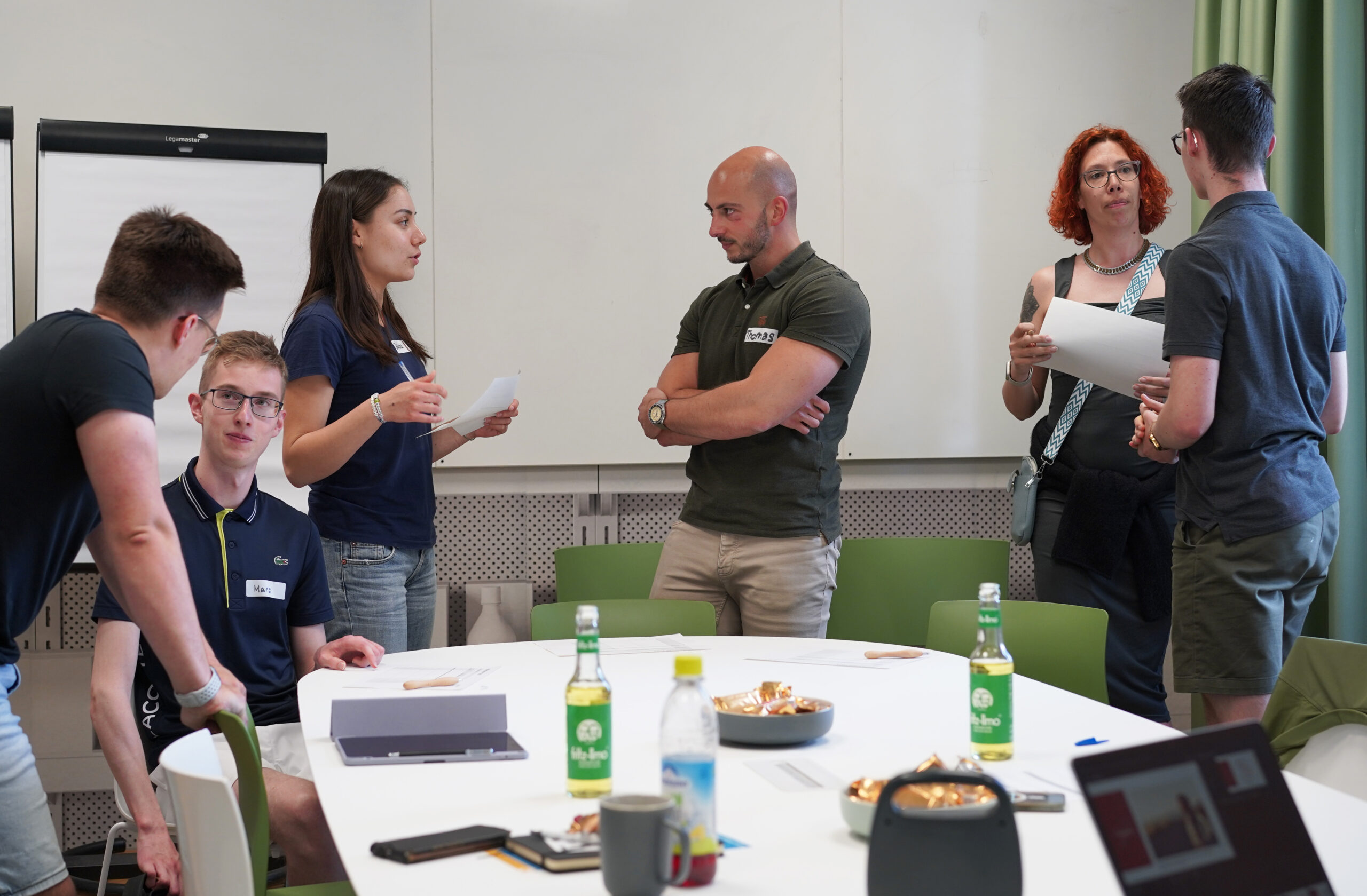 Eine Gruppe von Leuten der Hochschule Coburg diskutiert angeregt an einem Tisch mit Unterlagen und Snacks. Einige stehen, andere sitzen, im Hintergrund ein Flipchart. Verschiedene Getränke sind über den Tisch verteilt, alle bleiben konzentriert und beteiligen sich aktiv am Meeting.