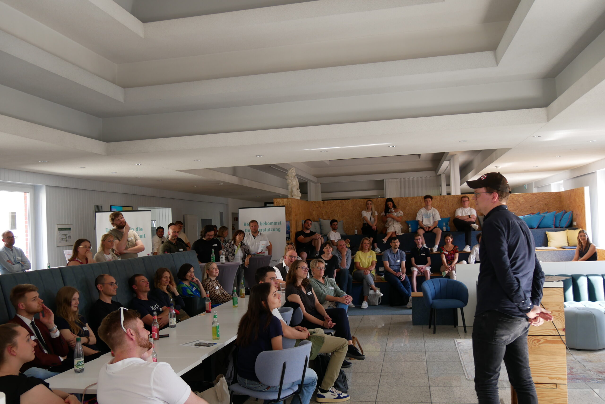 A group of people seated and standing in a modern, bright room at Hochschule Coburg attentively listens to a speaker at the front. The space boasts tiered seating, light-colored walls, and a high ceiling.