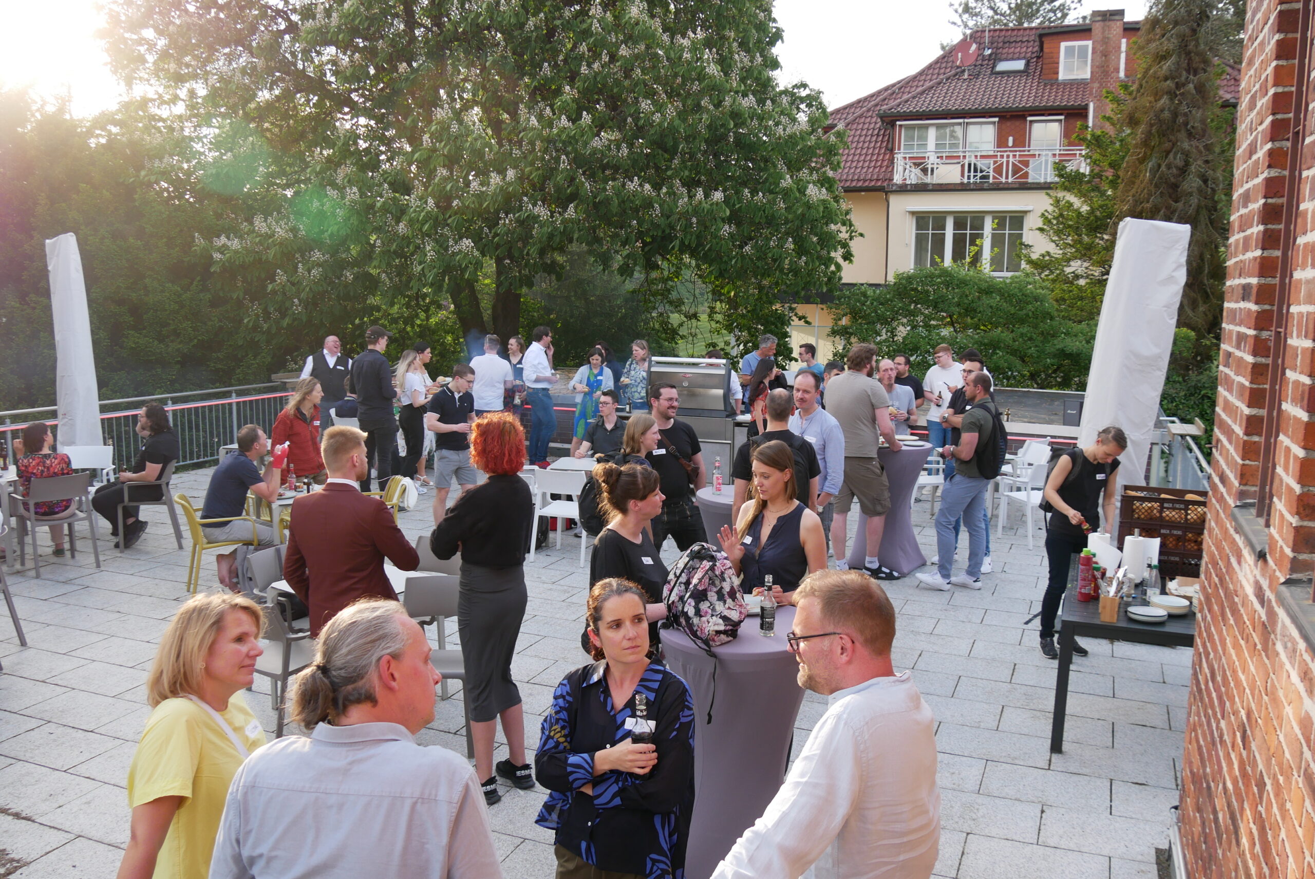 Eine große Gruppe von Menschen trifft sich bei einer Veranstaltung im Freien auf einer sonnigen Terrasse der Hochschule Coburg. Im Hintergrund stehen Bäume, Tische und Stühle verteilt und ein Backsteingebäude ist zu sehen. Die Menschen unterhalten sich und genießen das Beisammensein.