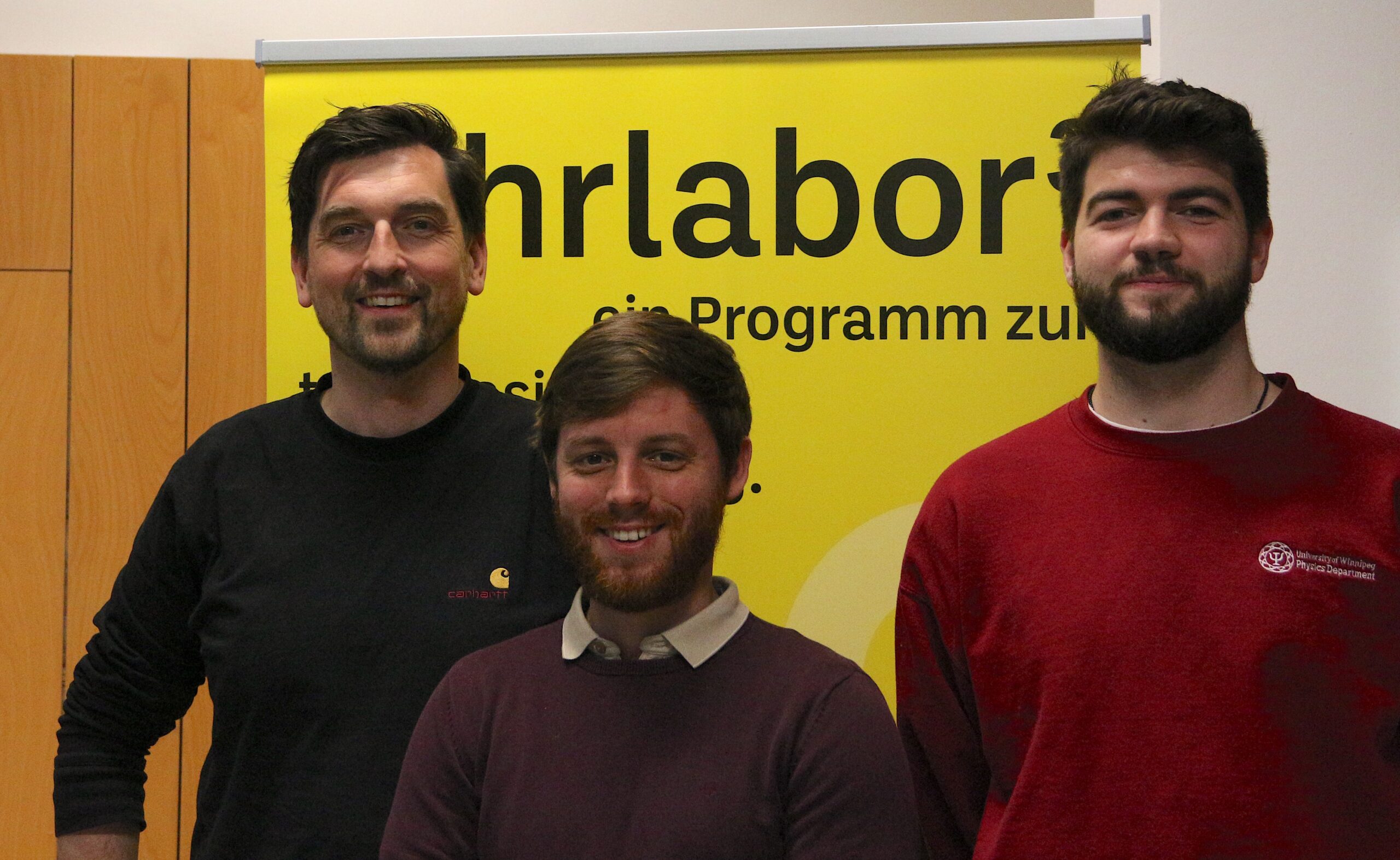 Three men stand smiling in front of a yellow banner with partial text visible inside a room adorned with wooden paneling. Casually dressed in sweaters, these individuals appear to be part of an event at Hochschule Coburg.