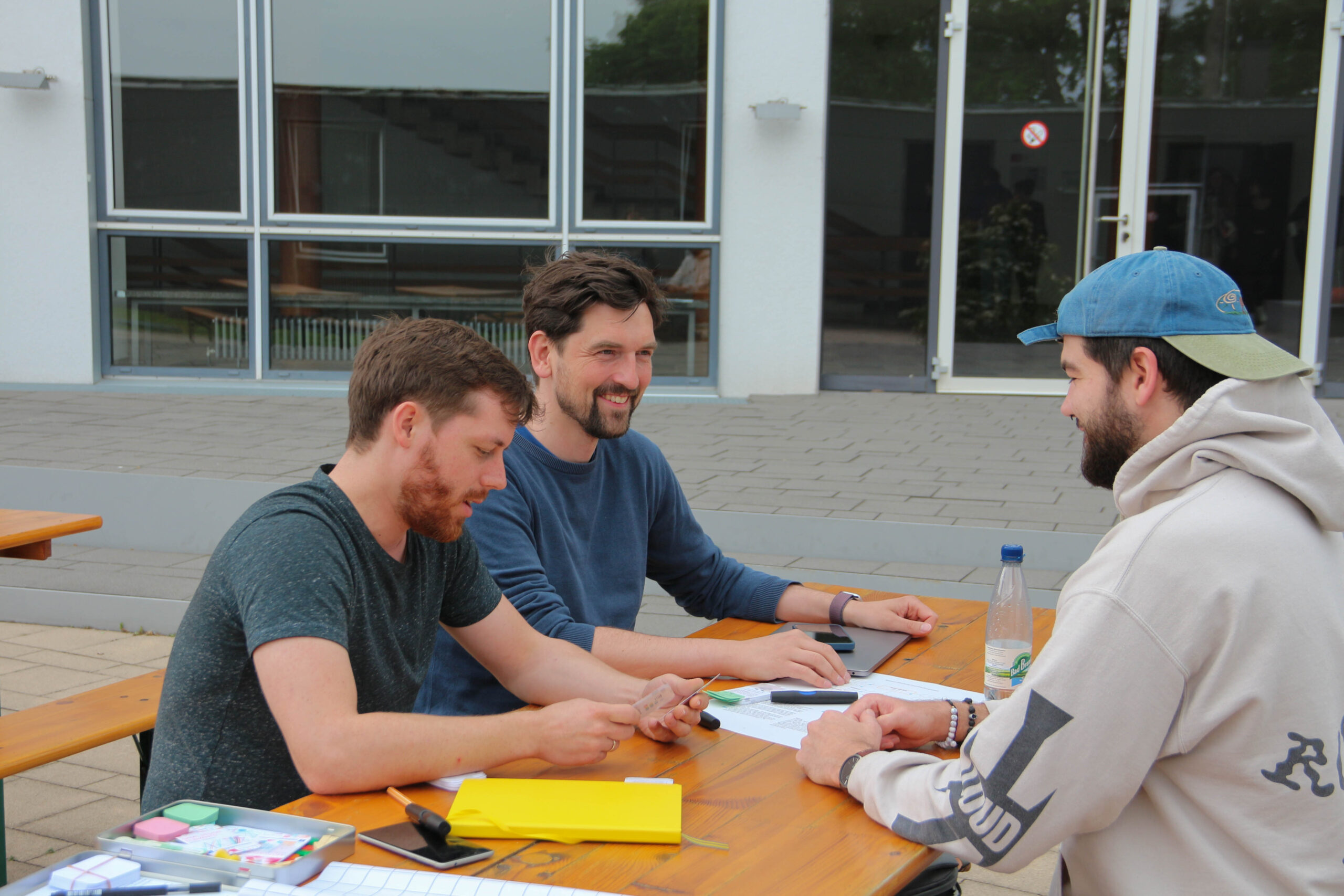 Three men are sitting at a wooden table outside Hochschule Coburg, engaged in conversation. They have a laptop, papers, and a yellow pencil case on the table. The background showcases a building adorned with glass windows.