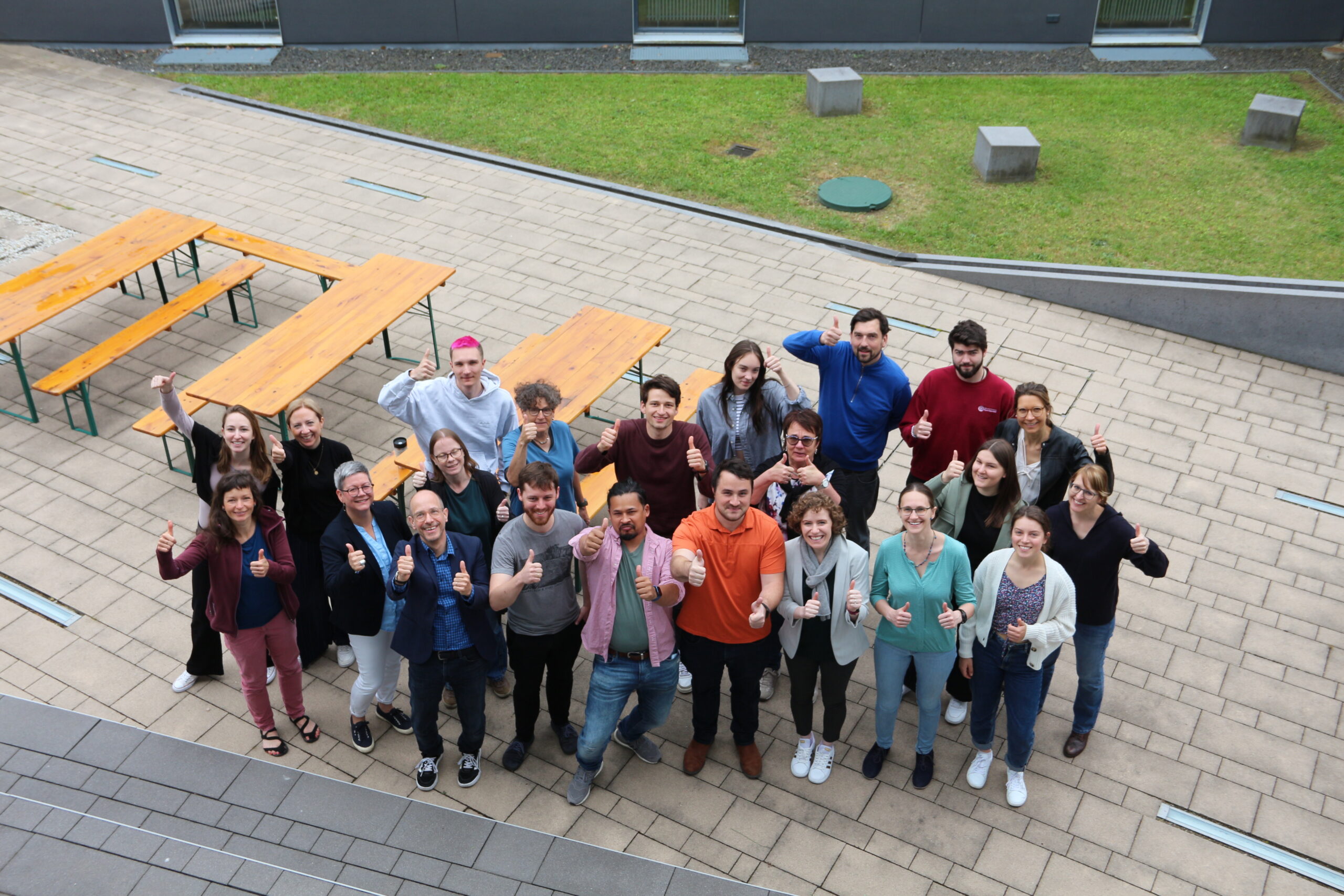 A group of people stand outdoors on a paved area with picnic tables at Hochschule Coburg, smiling and giving thumbs up. They are casually dressed, and greenery is visible in the background.