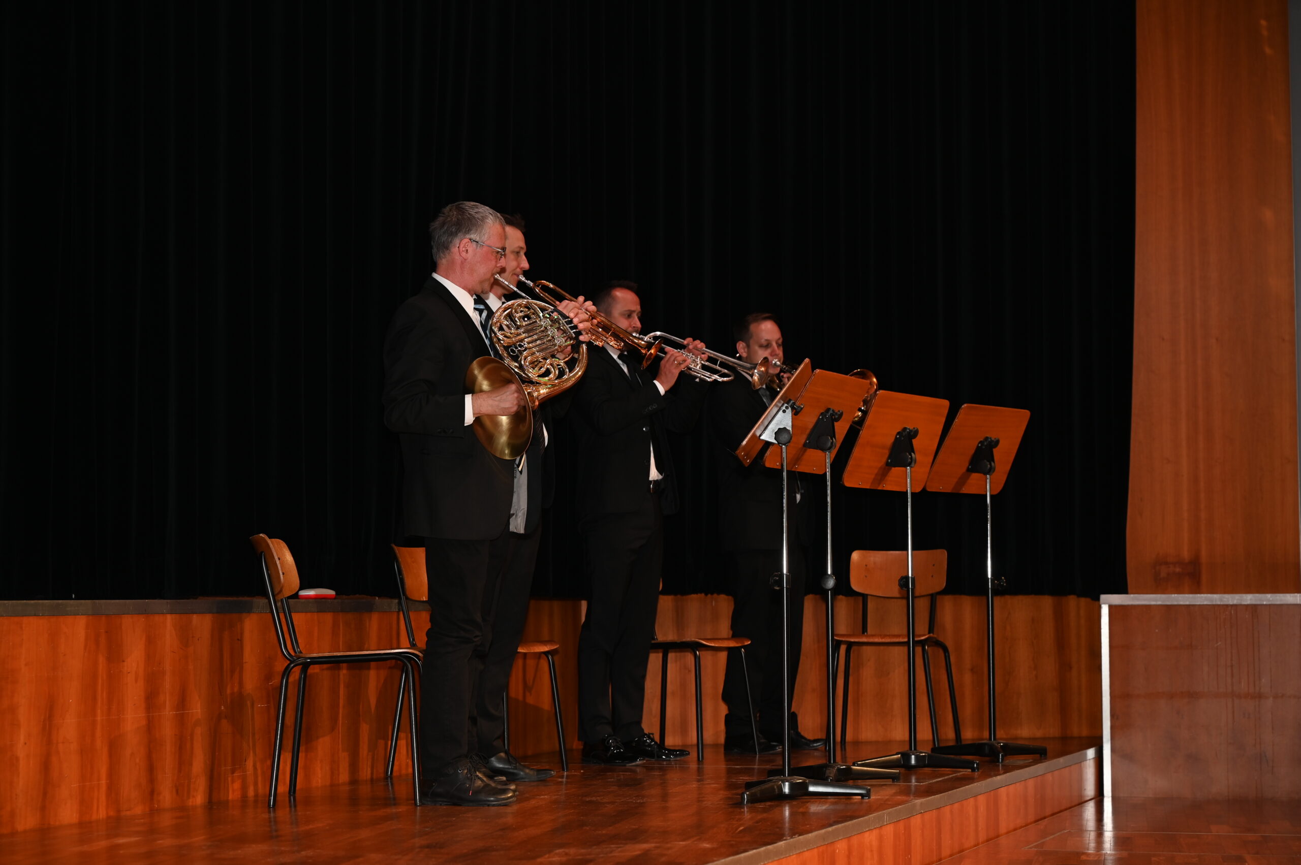 Drei Musiker in formellen Anzügen, die die Hochschule Coburg repräsentieren, treten mit Blechblasinstrumenten auf der Bühne auf. Zwei spielen Trompeten und einer Waldhorn. Sie stehen vor Notenständern mit Notenblättern auf einer Holzbühne, eingerahmt von einem dunklen Vorhanghintergrund.