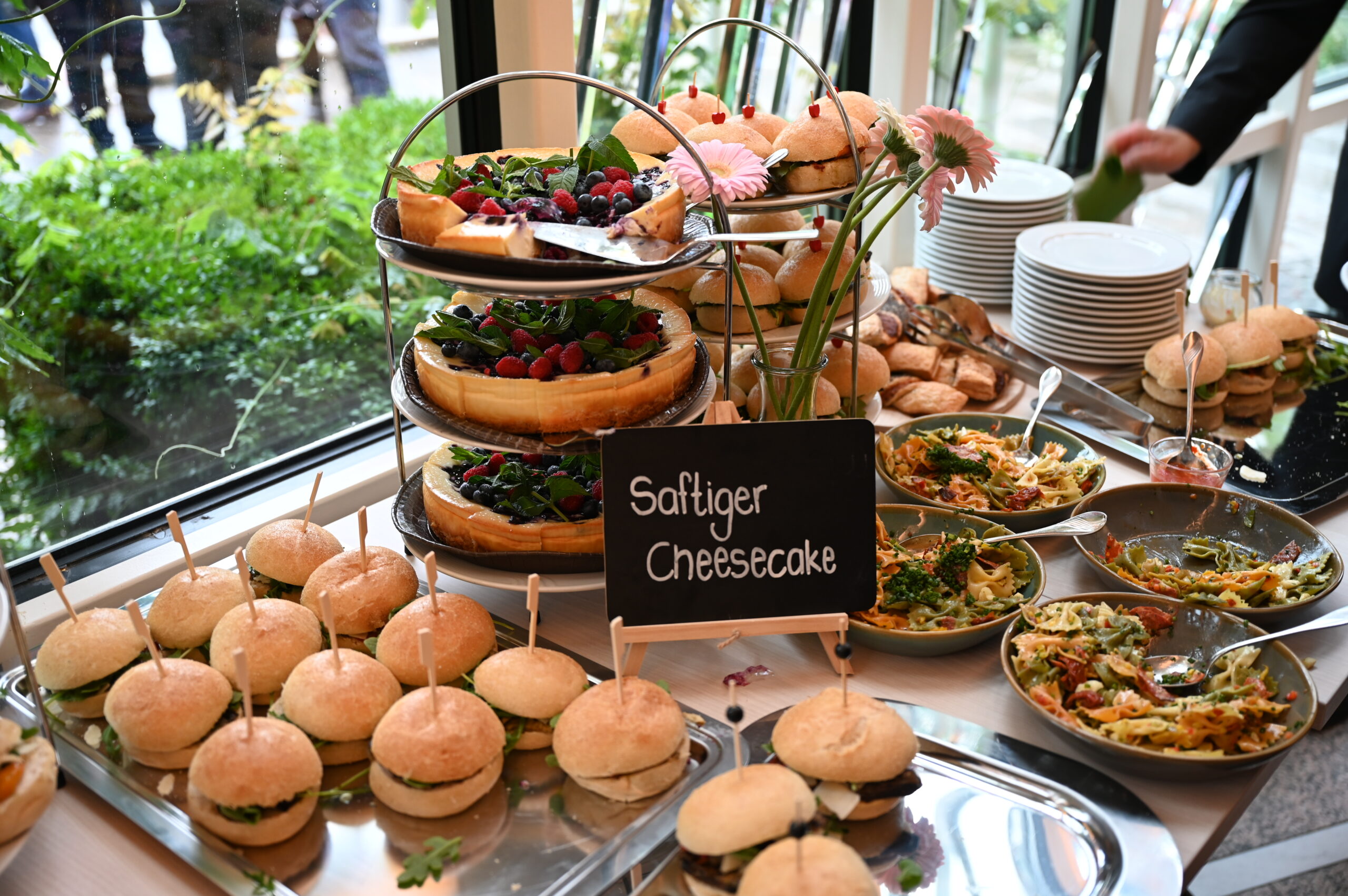 Ein Buffettisch voller Köstlichkeiten – Tabletts mit Mini-Sandwiches, Schüsseln mit Nudelsalat und eine Etagere mit Käsekuchen, gekrönt mit Beeren. Ein Kreideschild verkündet fröhlich „Saftiger Käsekuchen“. Teller und Besteck warten schon, vielleicht ein Hinweis auf eine Versammlung der Hochschule Coburg.