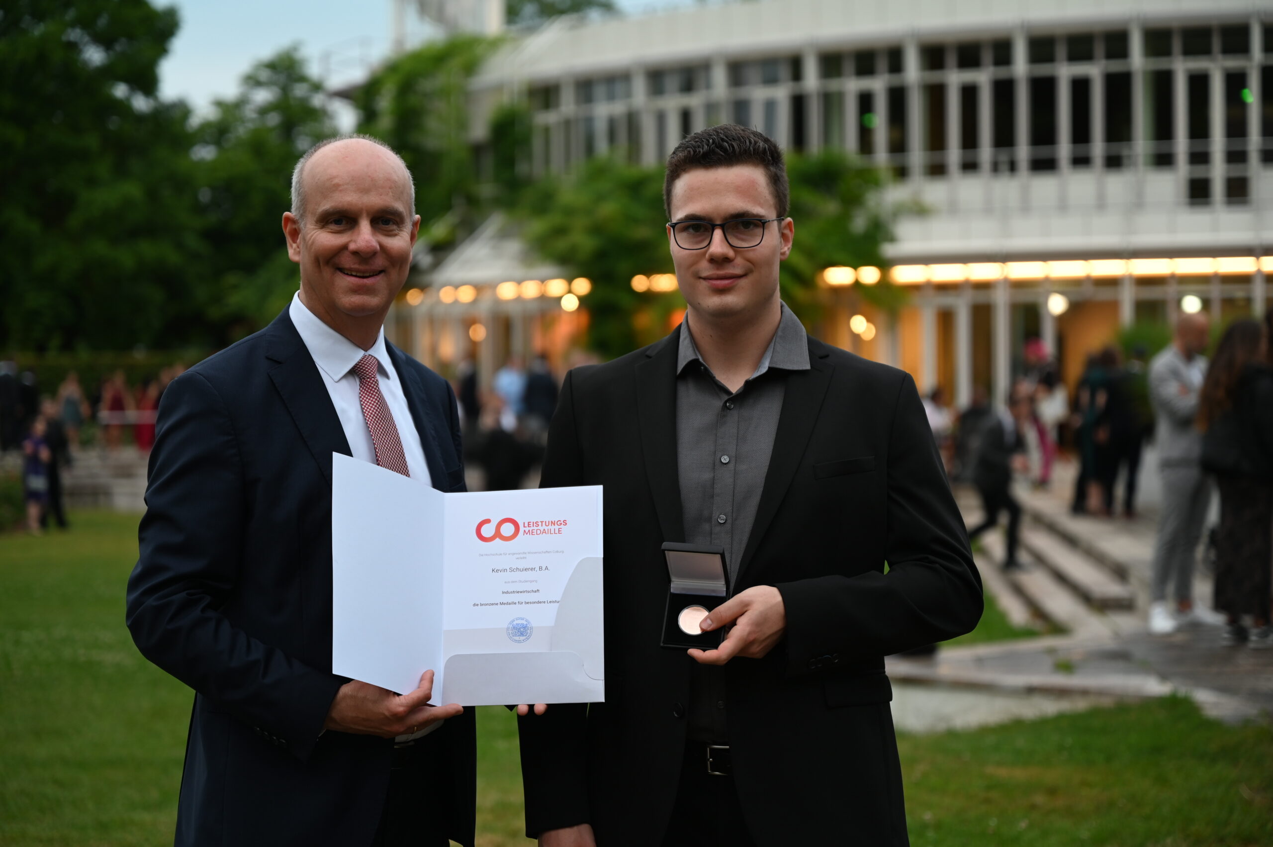 Zwei Männer in Anzügen stehen draußen vor der Hochschule Coburg. Der Mann links hält eine Urkunde mit dem Logo „GO“, während der Mann rechts einen kleinen runden Gegenstand in den Händen hält. Sie lächeln beide, im Hintergrund sind ein Gebäude und Menschen zu sehen.