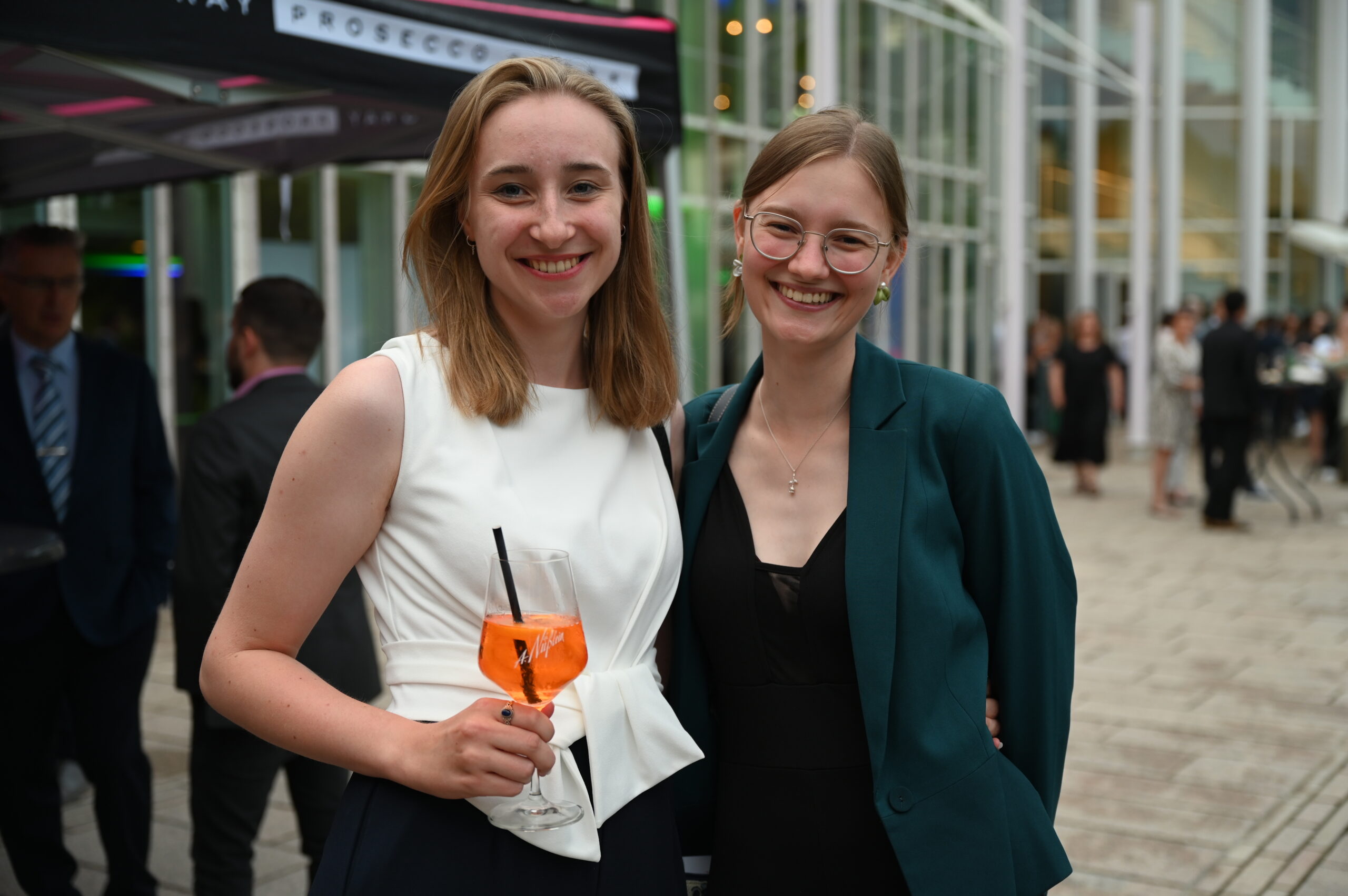 Zwei lächelnde Frauen bei einer Veranstaltung im Freien, die von der Hochschule Coburg veranstaltet wird. Eine hält ein Glas mit einem Orangengetränk. Sie sind in formelle Kleidung gekleidet, im Hintergrund ist moderne Architektur zu sehen.