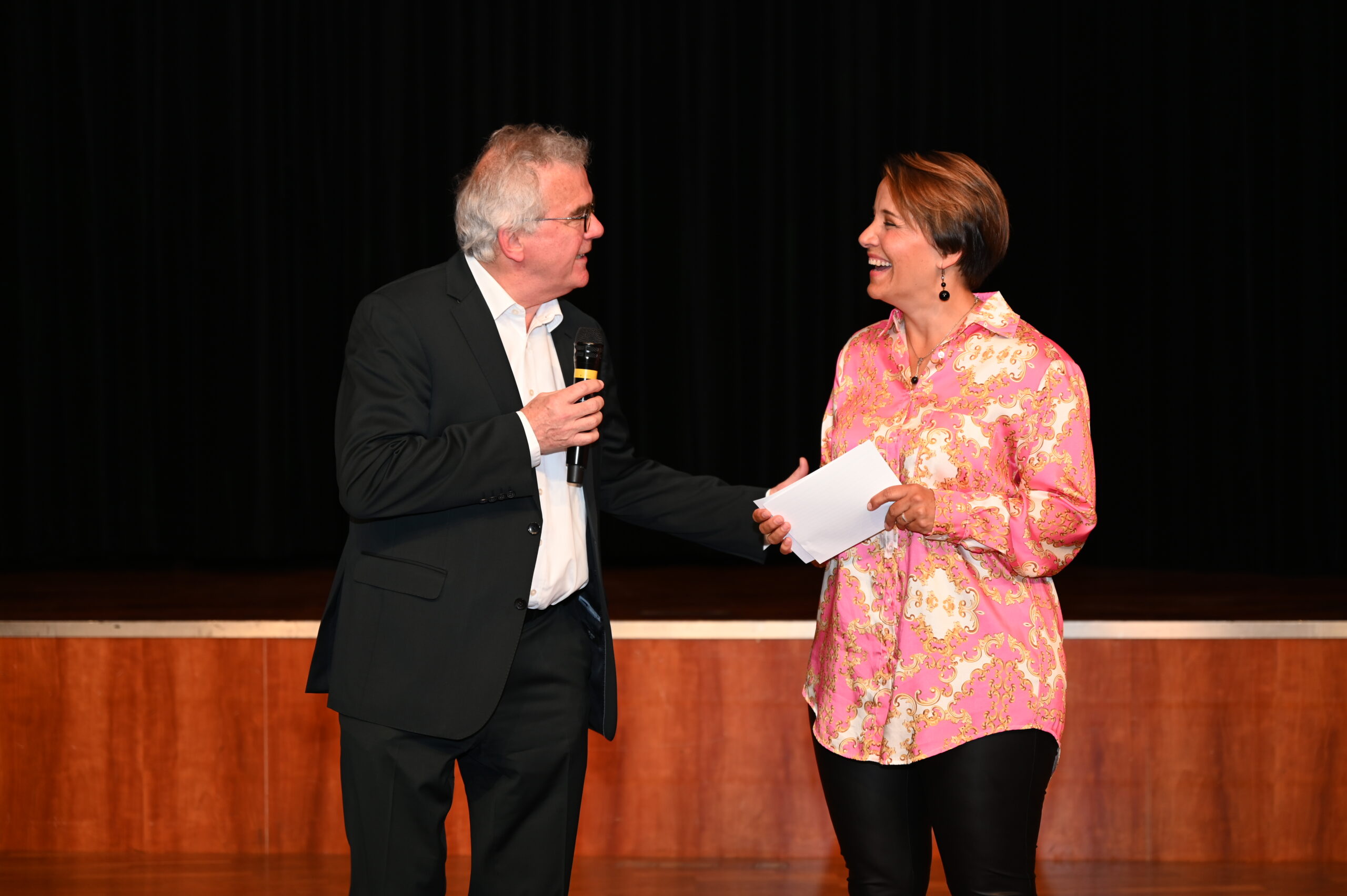 Ein Mann im Anzug hält ein Mikrofon in der Hand und unterhält sich herzlich mit einer Frau in einer rosa geblümten Bluse, die einen Umschlag hält. Sie stehen auf der Holzbühne der Hochschule Coburg, eingerahmt von einem schwarzen Vorhang, und lächeln sich an.