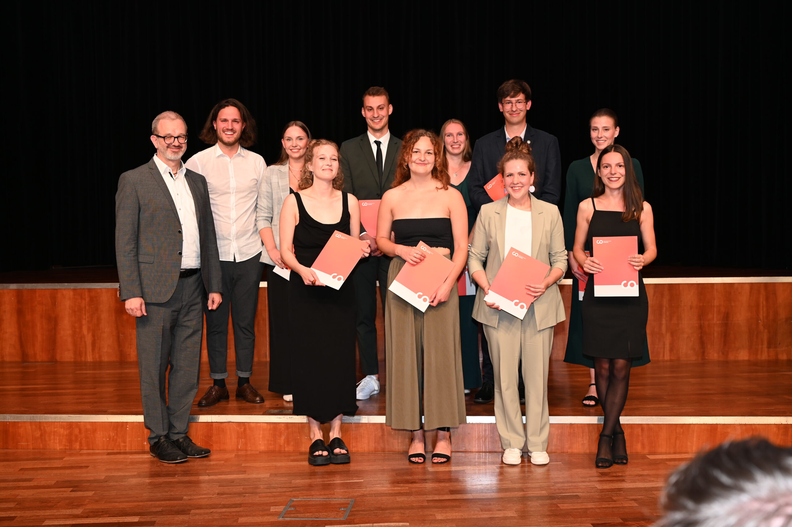 Eine Gruppe von elf Personen steht lächelnd auf einer Bühne und repräsentiert voller Stolz die Hochschule Coburg. Einige halten orangefarbene Mappen in den Händen. In formeller Kleidung stehen sie vor einem dunklen Vorhang und positionieren sich anmutig in zwei Reihen auf dem polierten Holzboden.