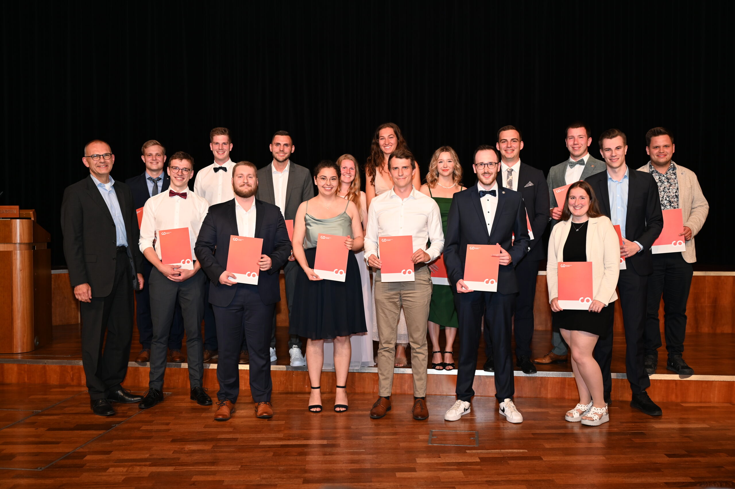 Eine Gruppe von Menschen steht mit Zertifikaten in den Händen auf der Bühne der Hochschule Coburg und lächelt. Sie sind in formelle Kleidung gekleidet, im Hintergrund ein dunkler Vorhang.