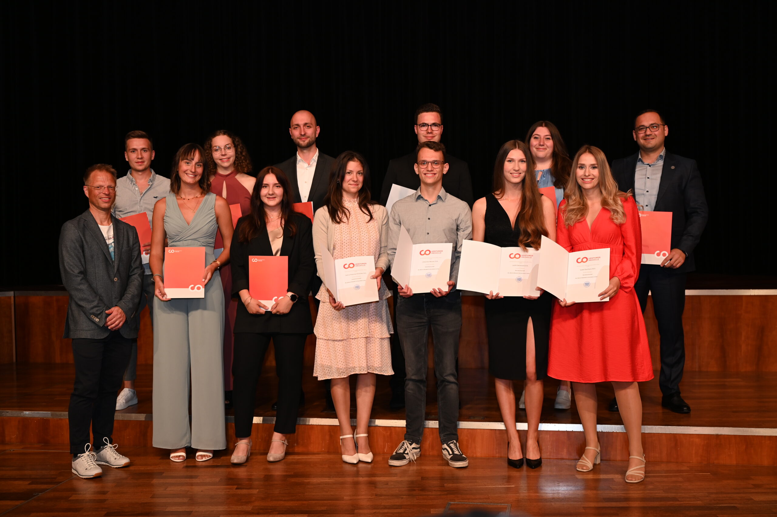 Eine Gruppe von 13 Personen der Hochschule Coburg, halbformell gekleidet, steht mit Zertifikaten und Mappen auf einer Bühne. Ihre Lächeln vor dem Hintergrund eines dunklen Vorhangs lassen auf eine Feier oder Preisverleihung schließen.